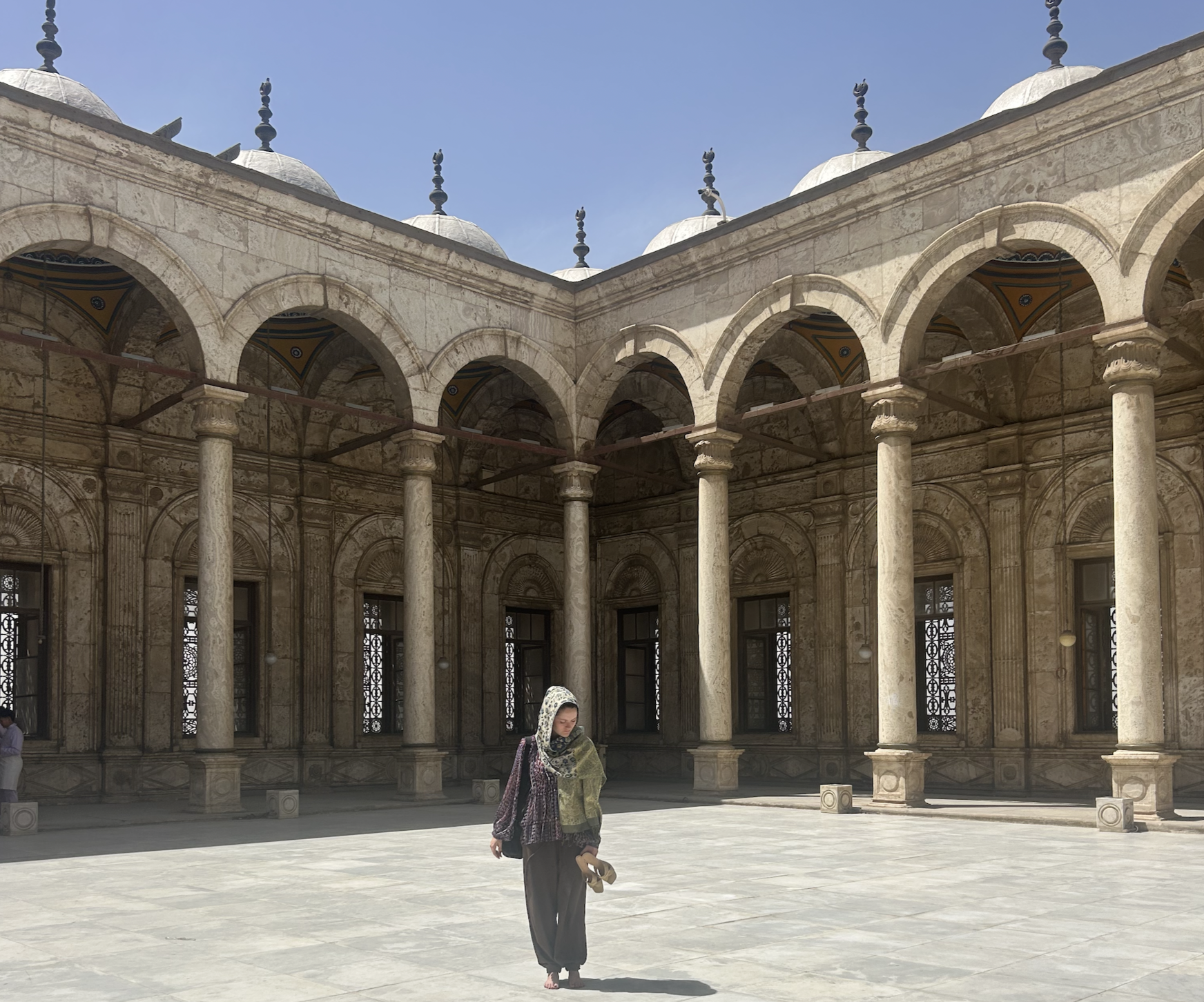 A woman in traditional clothing and headscarf walks barefoot across a courtyard with a stone arcade featuring arches and columns, under a clear blue sky.