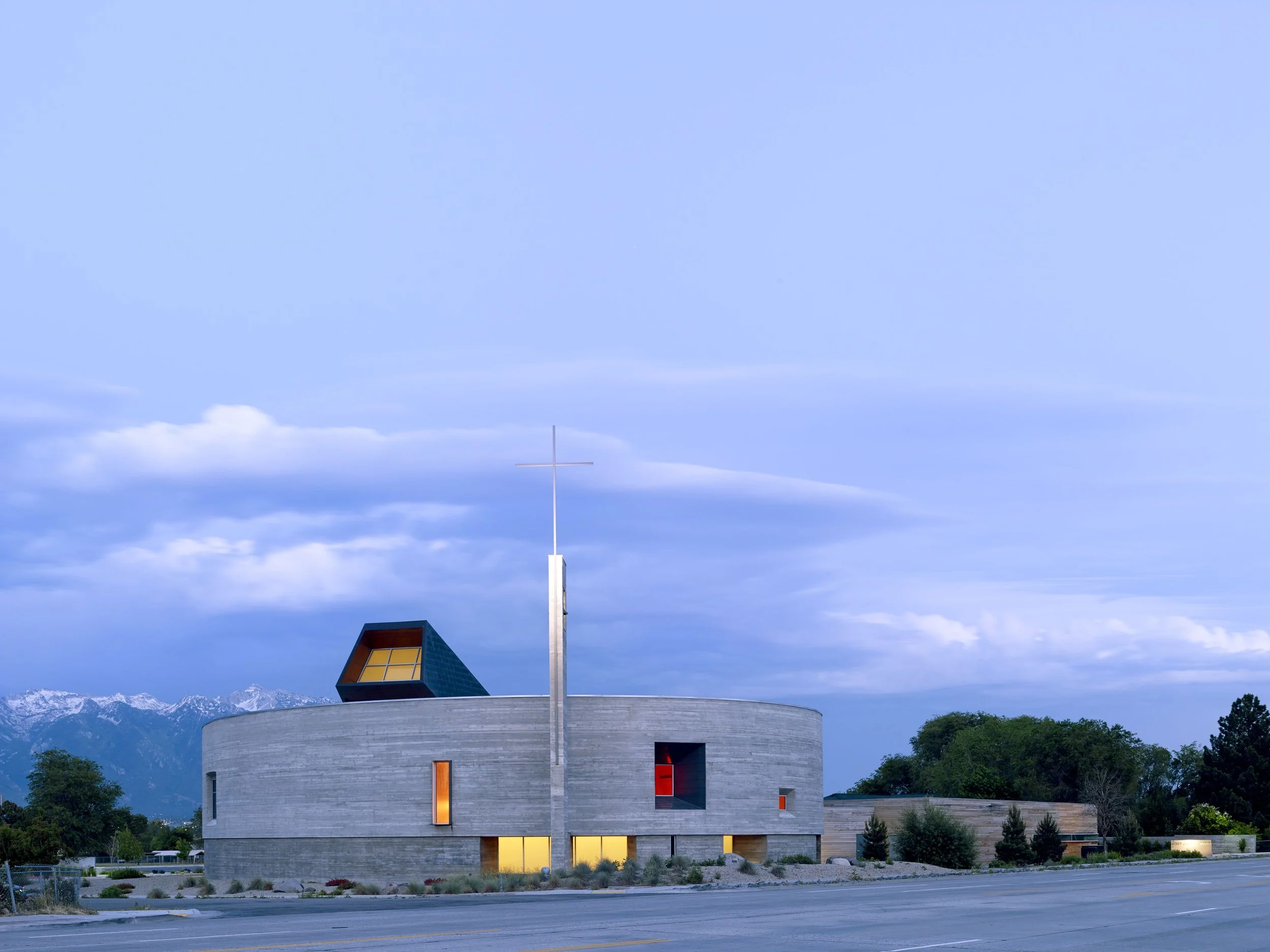Modern church building with a circular concrete structure, large windows, a cross on a tall pole, and a mountains backdrop under a cloudy sky.