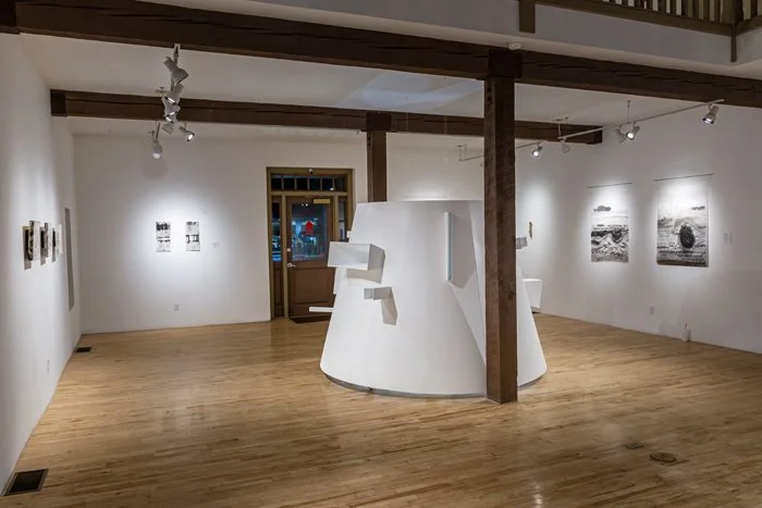 An art gallery featuring black-and-white photographs on the walls and a white sculptural installation in the center of the room, with wooden beams and hardwood floors.