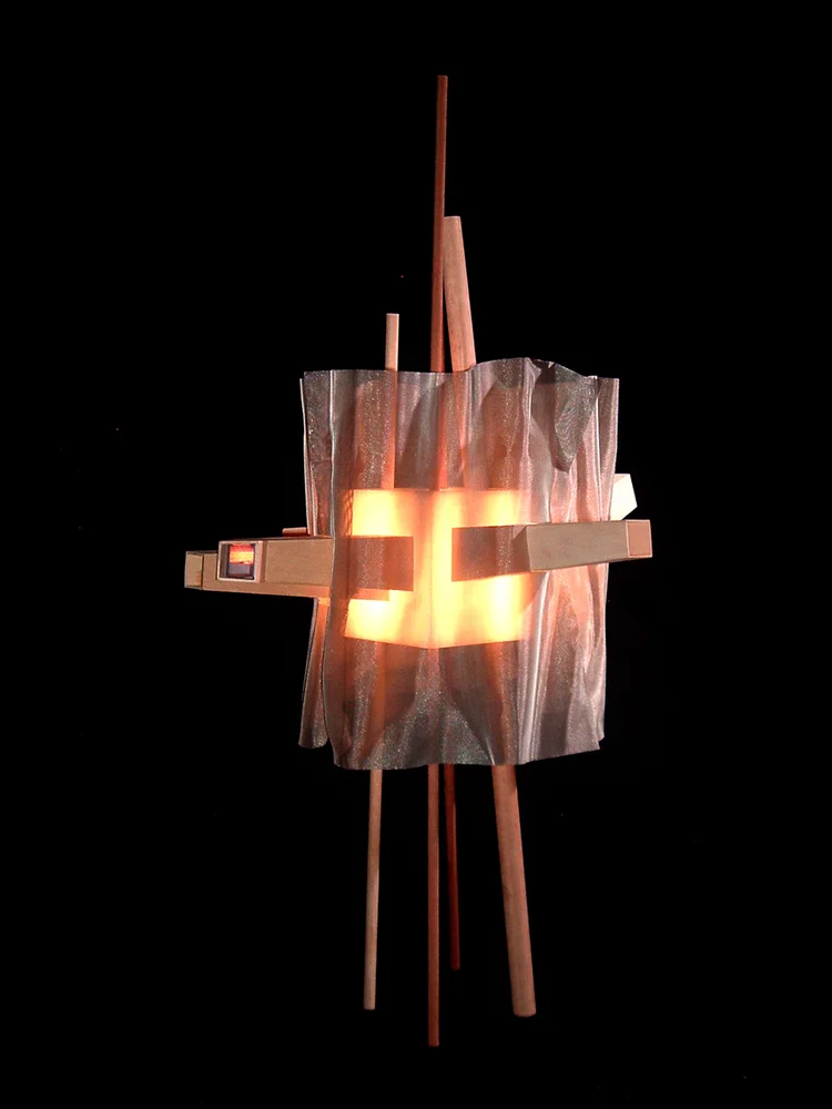 Artistic sculpture of a face with glowing eyes and a metallic surface, mounted on wooden sticks against a black background.