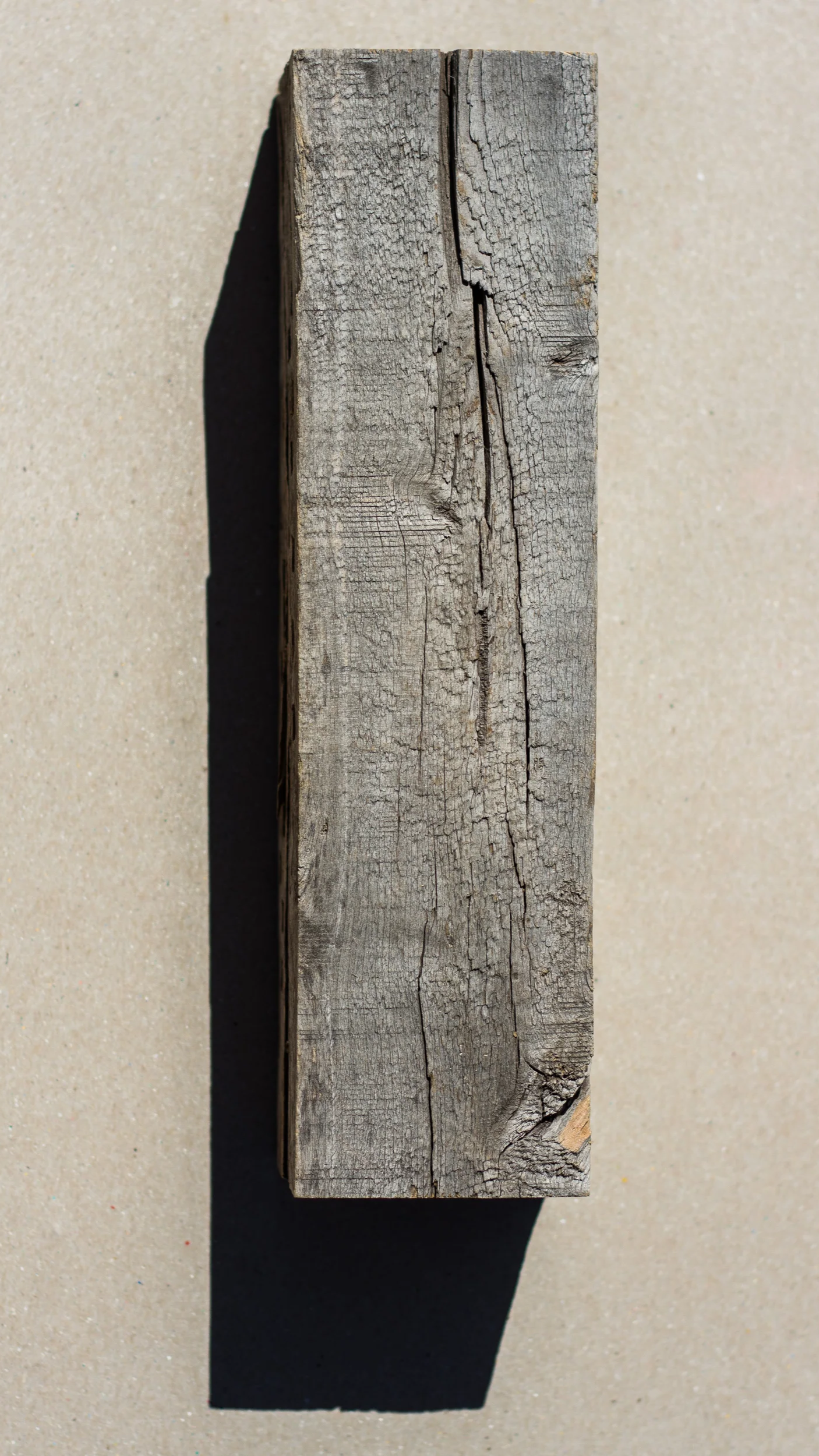A weathered, rectangular piece of wood with visible cracks and grain, casting a shadow on a beige sandy background.