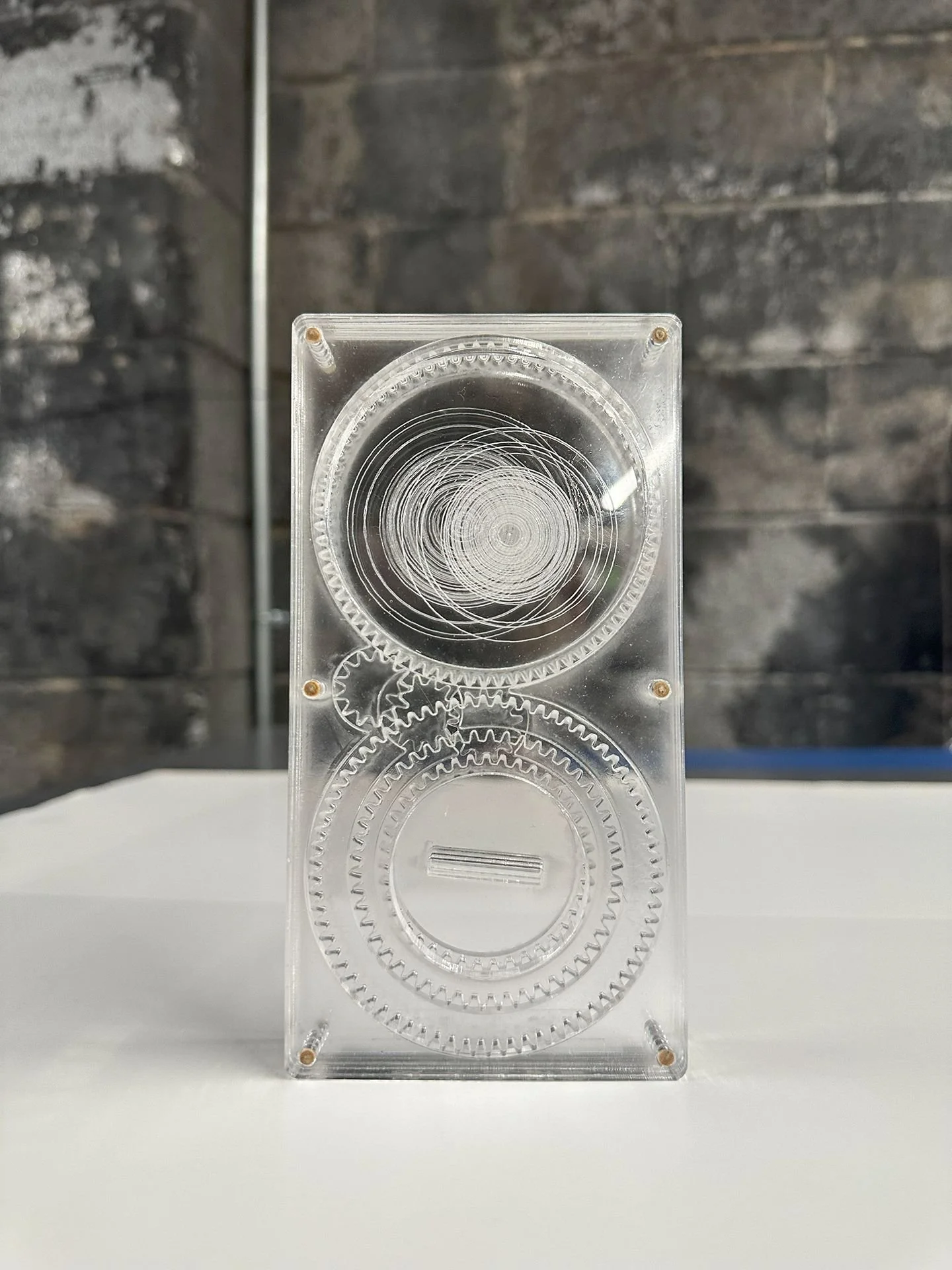 Top view of a clear rectangular plastic puzzle box with visible interlocking gears and mechanisms inside, placed on a white surface against a dark textured background.
