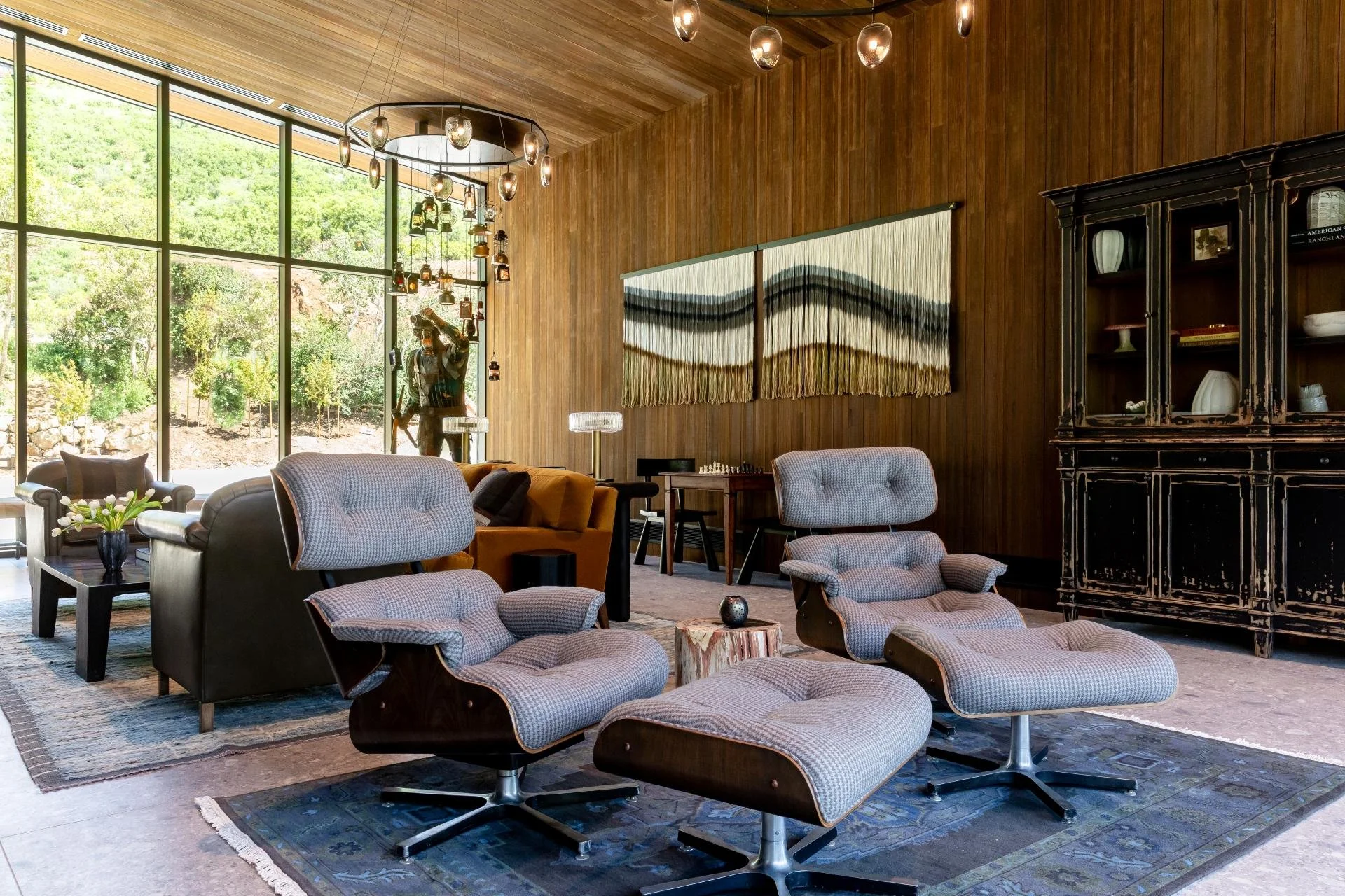 Modern living room with large glass windows, wooden walls, corner black cabinet, two armchairs with matching footstools, yellow sofa, small tables, decorative hanging lights, artwork on wall, and a garden view outside.