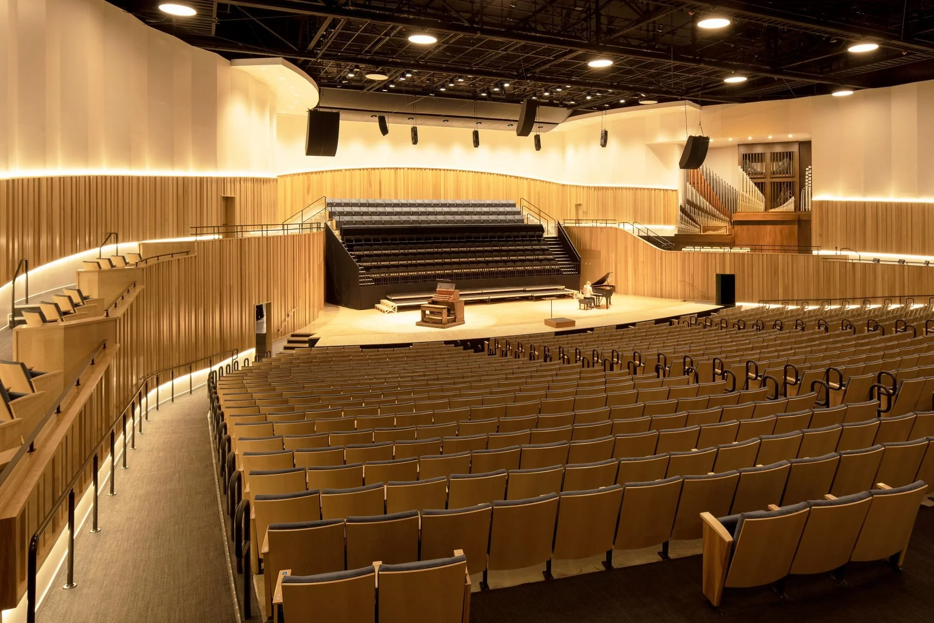 An empty concert hall with a stage featuring a grand piano and a few chairs, surrounded by warm wood paneling and modern lighting.