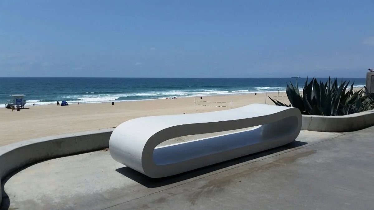 Modern white curved bench facing the beach with a view of the ocean, sandy shoreline, and blue sky.