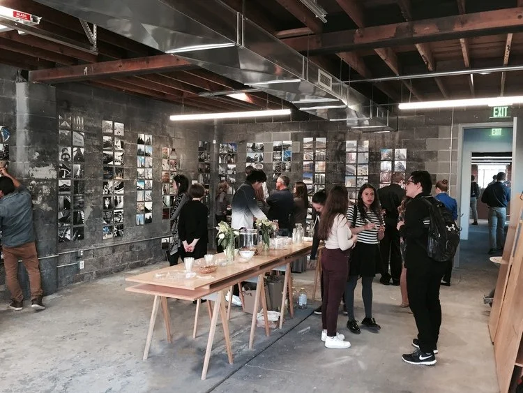 People mingling in an industrial-style art gallery with photo displays on the wall, a wooden table with snacks and drinks in the foreground, and an exit door to the right.