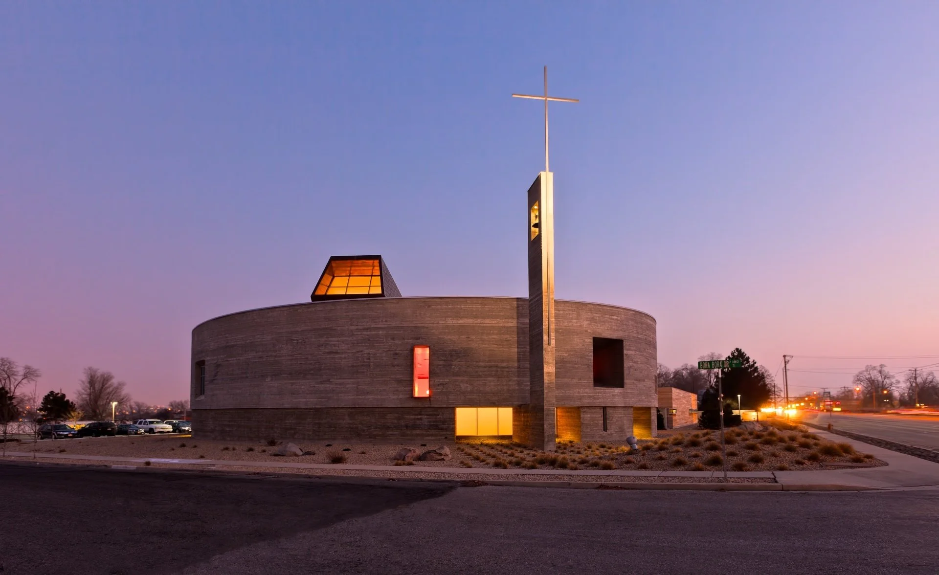 Boardform concrete church exterior glowing in warm sunset light with tall steel cross designed by Sparano + Mooney Architecture.