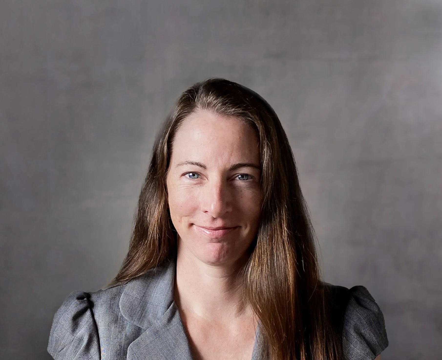 Portrait of a woman with long brown hair, blue eyes, and fair skin wearing a gray suit jacket, smiling slightly against a plain gray background.