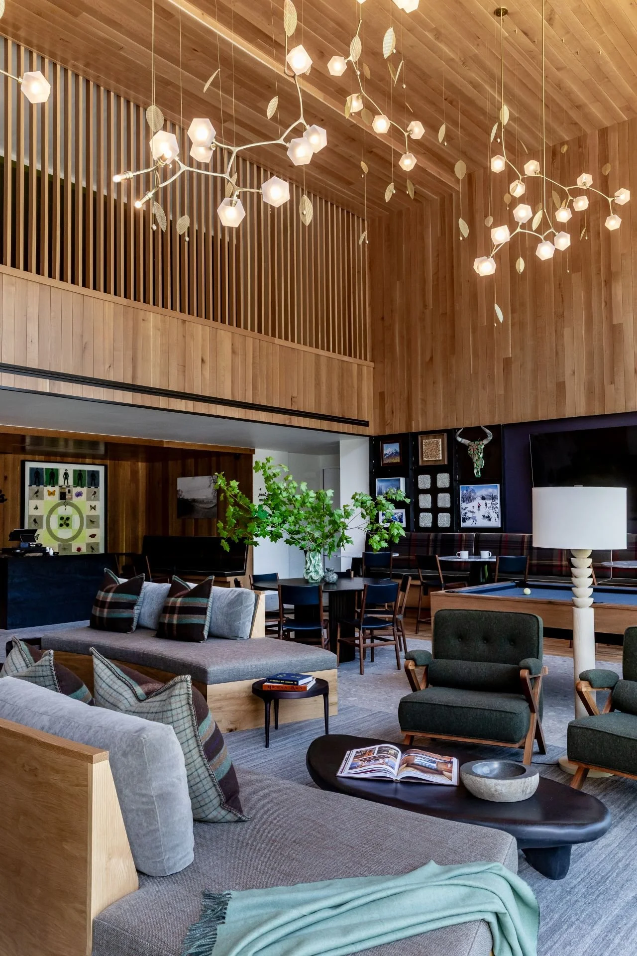 Spacious living room with wooden walls and ceiling, modern lighting fixture, and various seating arrangements including sofas and chairs, decorated with pillows and a blanket, with artwork on the walls and a large flat-screen TV.