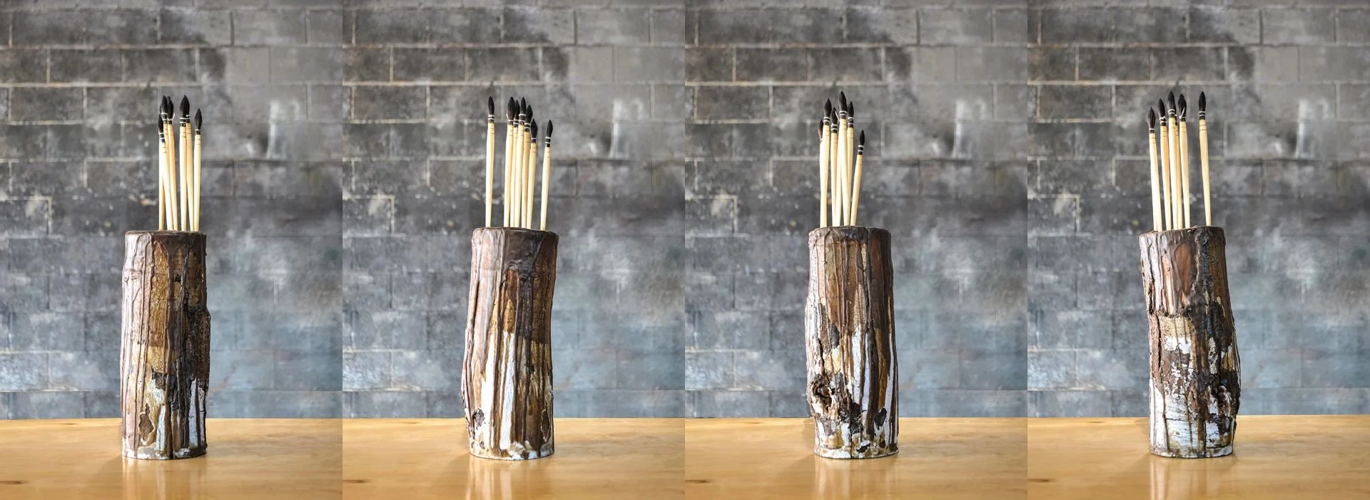 Four wooden vases with holes holding stick-like objects with black tips, placed on a wooden surface against a gray brick wall.