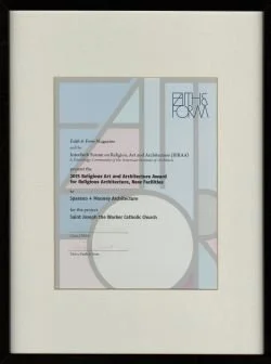 A framed certificate or award on a wall, with text indicating it is from the 2016 Religious Art and Architecture Award, presented by the Catholic Church for a project.