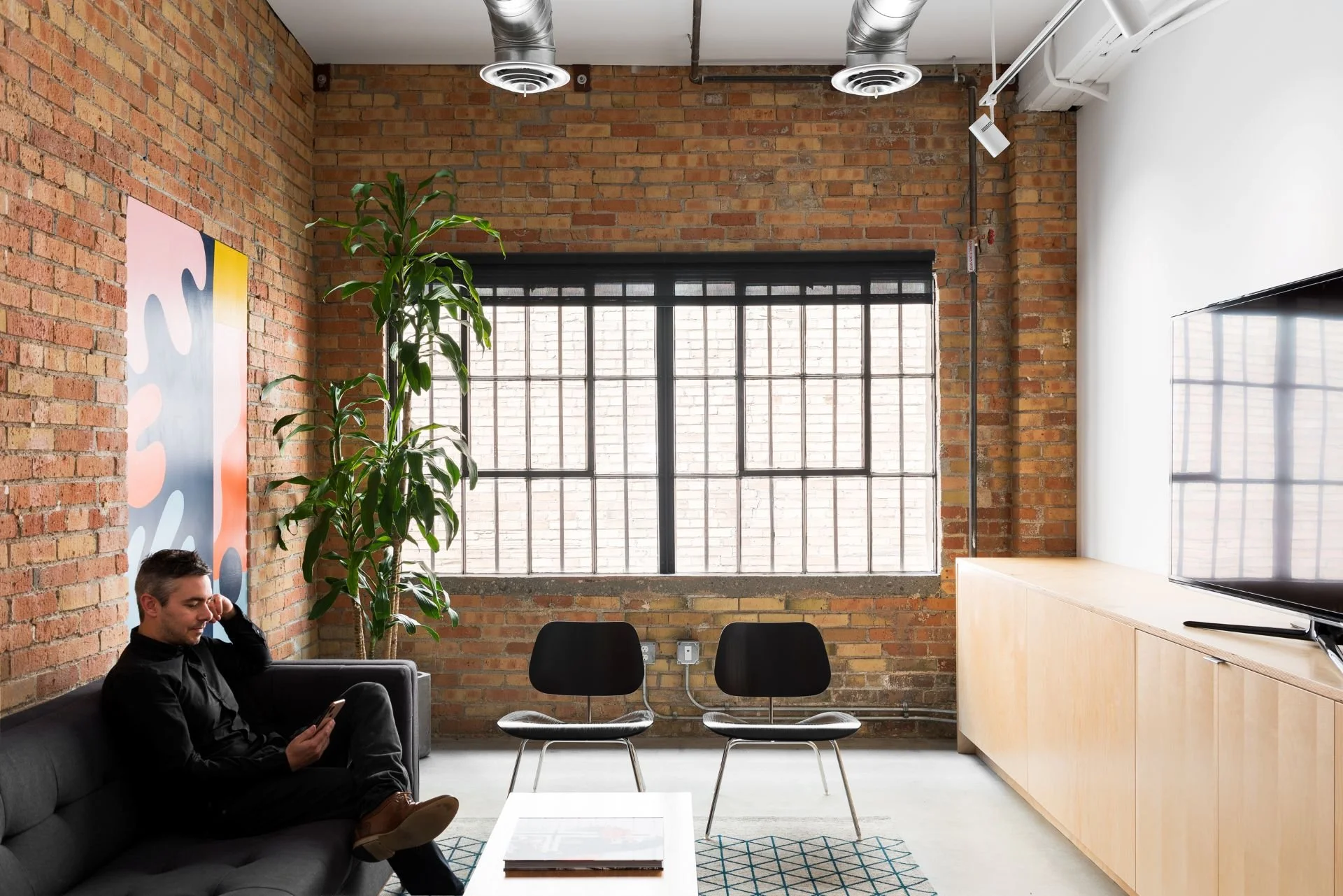 Modern office space with exposed brick walls, large industrial window, a man sitting on a gray couch using a phone, a tall plant next to the window, two black chairs, a wooden cabinet, a white coffee table with magazines, and abstract wall art.