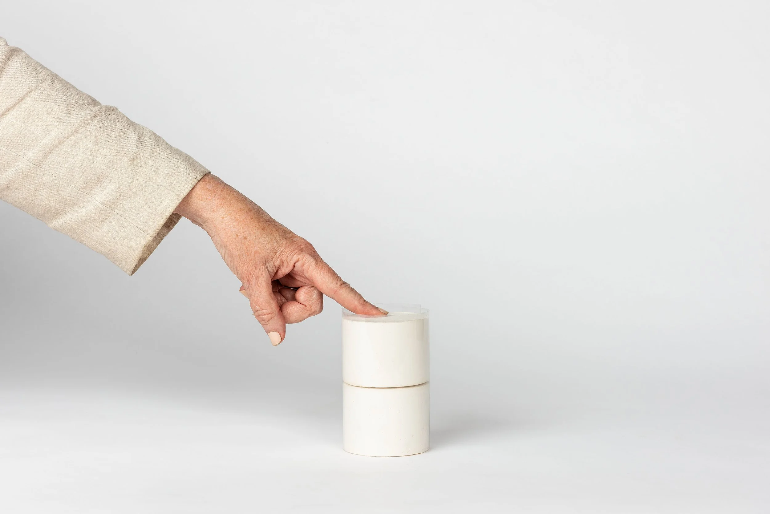Person's hand with a beige long sleeve shirt pointing at a white object on a plain white surface with a white background.