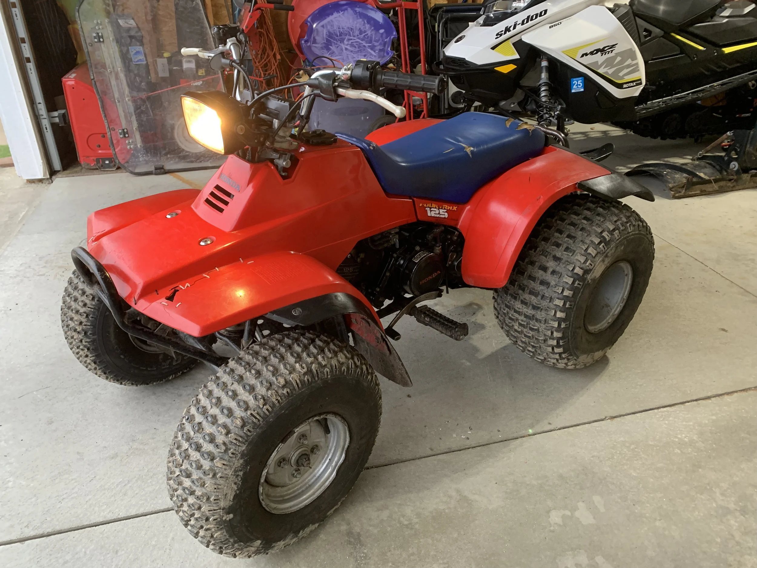 A red Honda four-wheeler ATV with knobby tires, parked indoors on a garage floor, next to other vehicles and equipment.