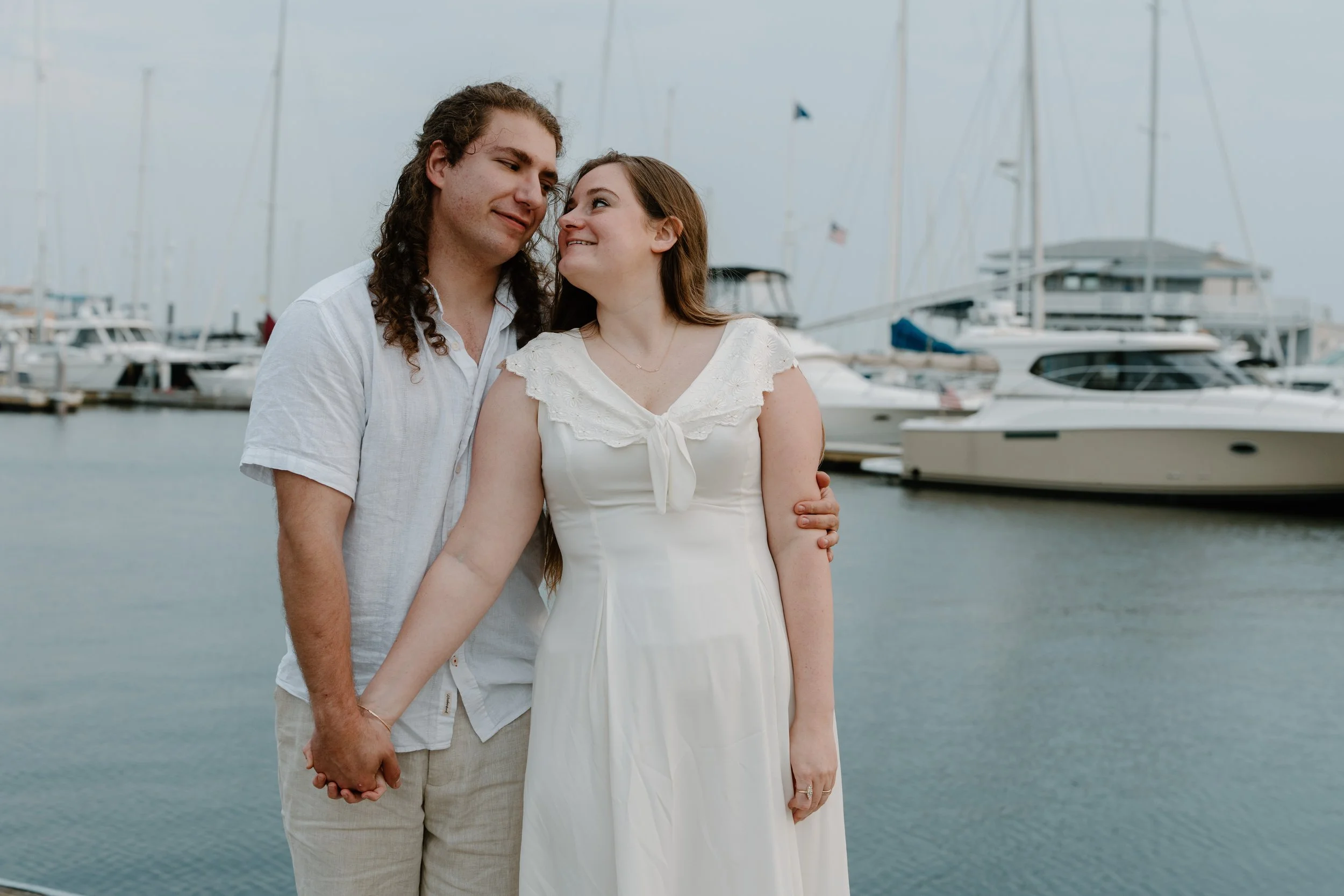 A couple holding hands by the marina, with sailboats and yachts in the background after an elopement in Newport, Rhode Island. 