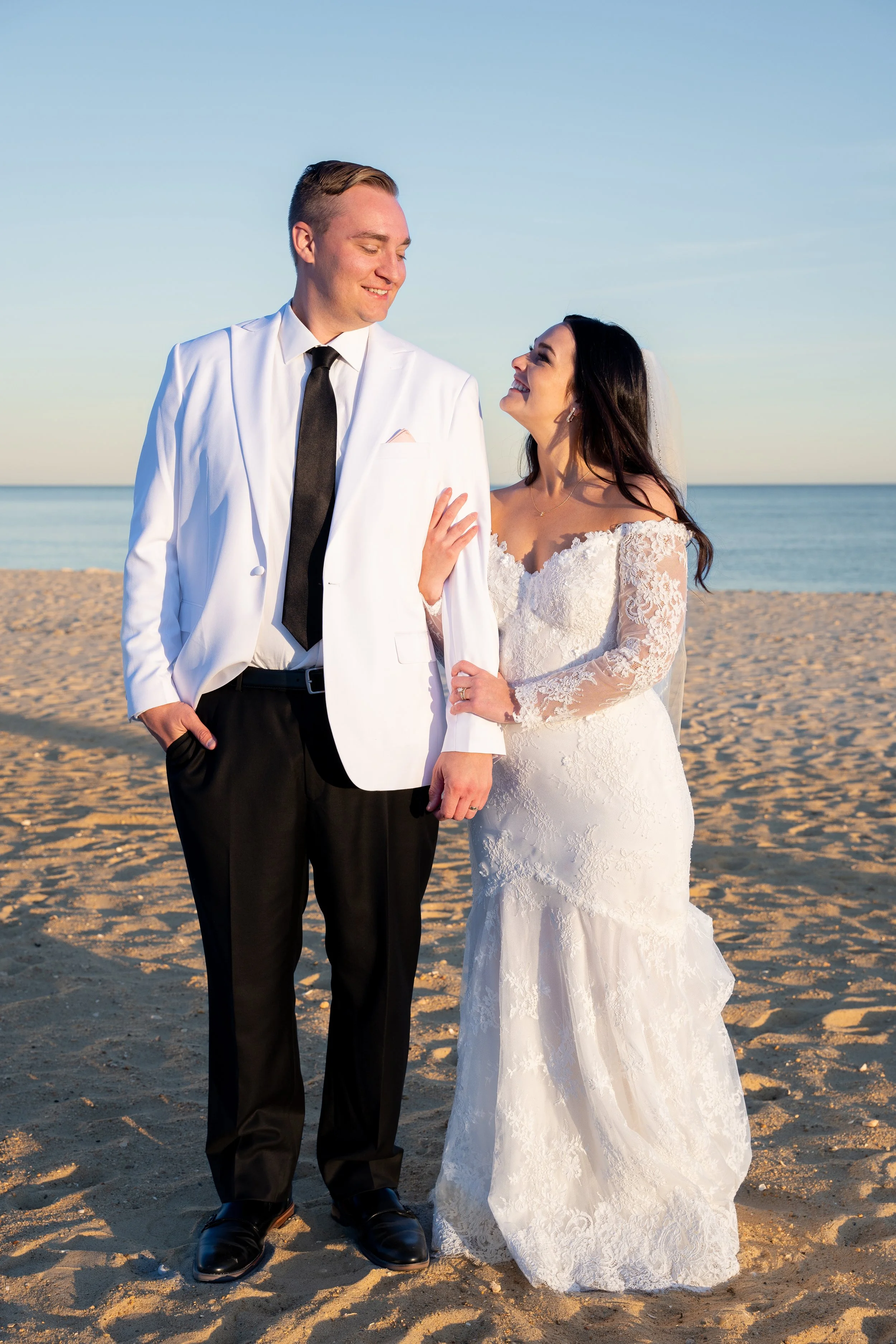 A wedding couple on the beach, the groom in a white tuxedo jacket and black trousers, and the bride in a lace wedding gown and veil, smiling at each other during sunset during a wedding at Red Jacket Resort in Cape Cod. 