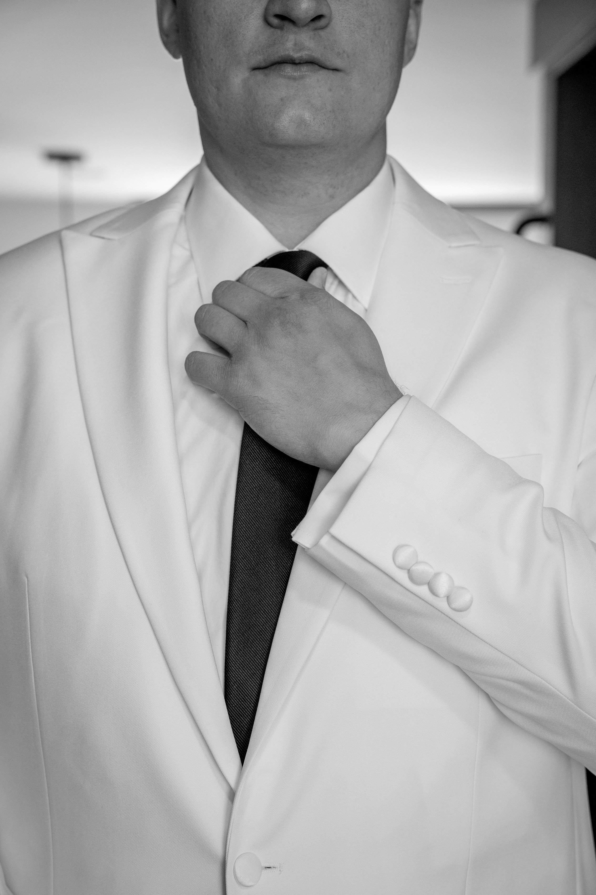 Close-up of a man in a white suit adjusting his black tie, with focus on his hand and suit during a wedding at Red Jacket Resort in Cape Cod. 