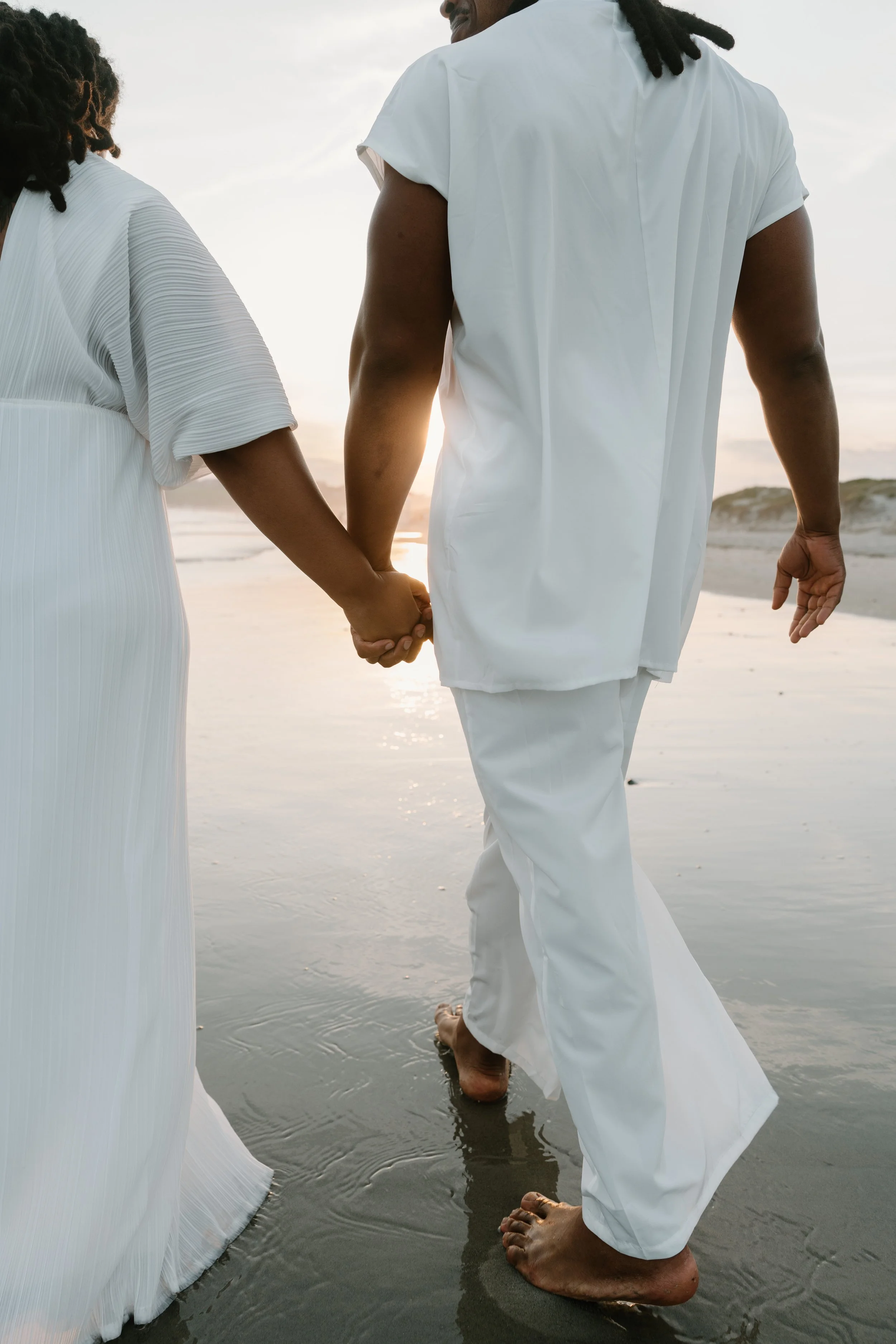 A couple holding hands walking on the beach during sunset, dressed in white clothing at an elopement at Second Beach in Rhode Island. 