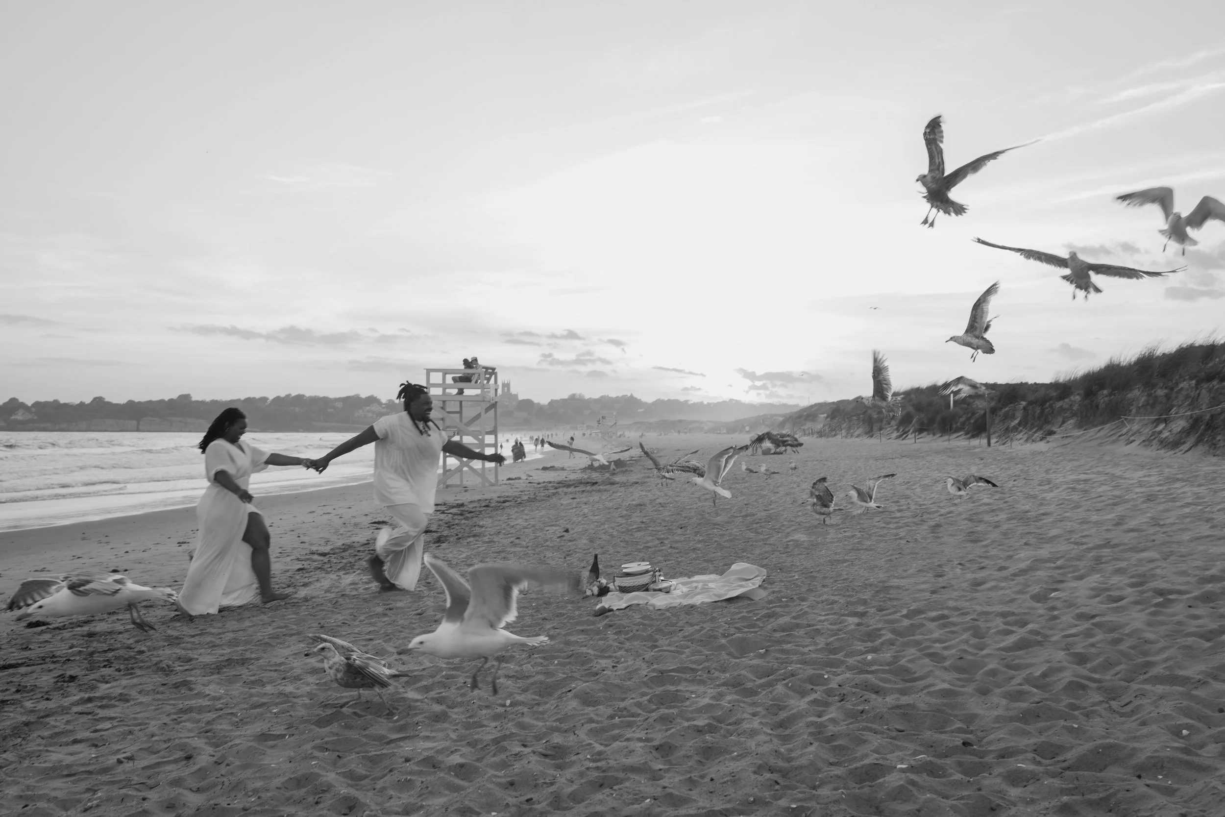 Two people holding hands on a beach with seagulls flying around them, a lifeguard tower in the background, and the sun setting in the sky at an elopement at Second Beach in Rhode Island. 