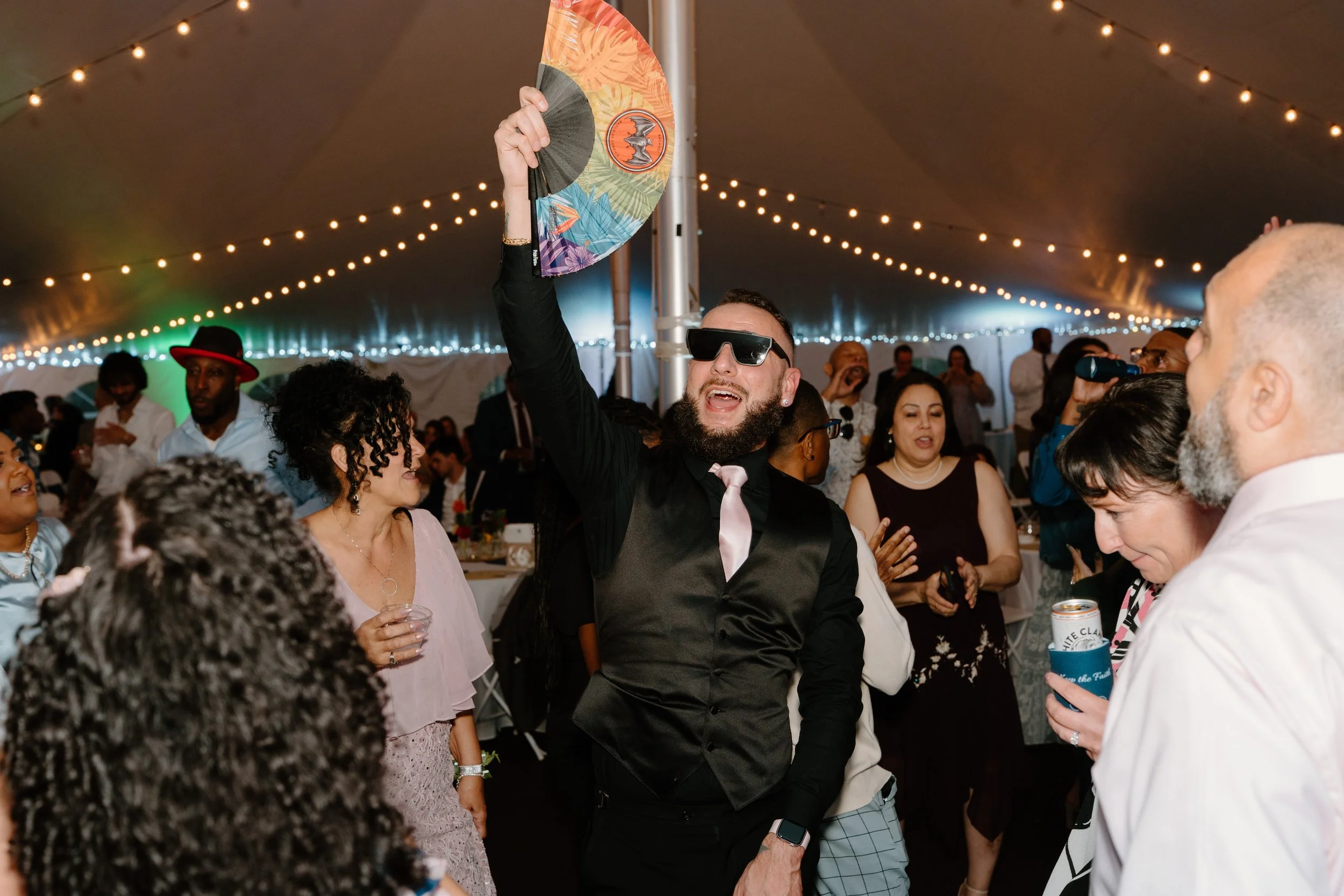 A group of people dancing and socializing under a large tent with string lights. One man wearing sunglasses, a black shirt, and a satin tie is holding a colorful fan and singing or shouting for a wedding at Holiday Hill Day Camp in Connecticut.