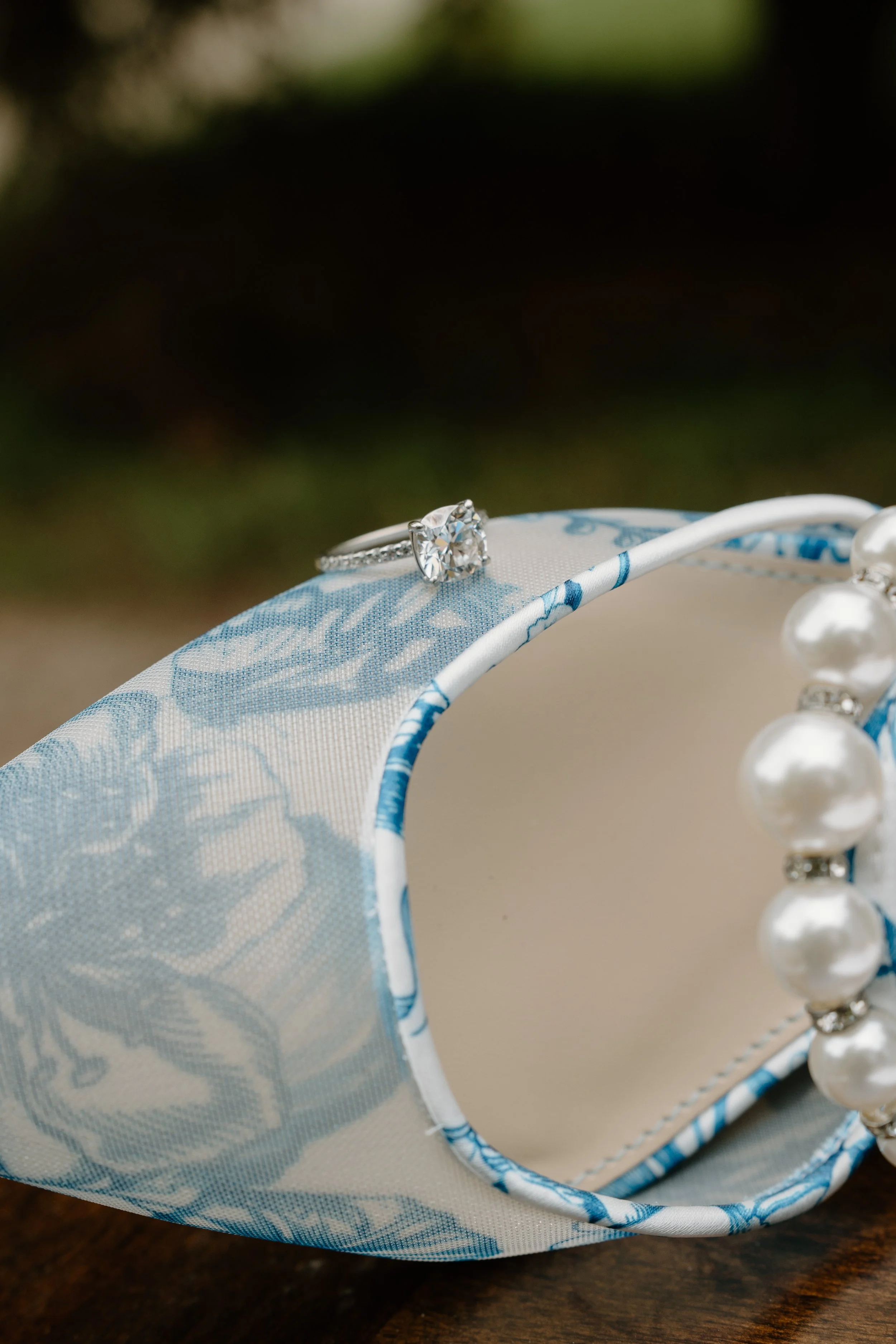 Close-up of a diamond engagement ring placed on a fabric-covered mirror with pearl necklace, on a wooden surface for a wedding at Holiday Hill Day Camp in Connecticut.