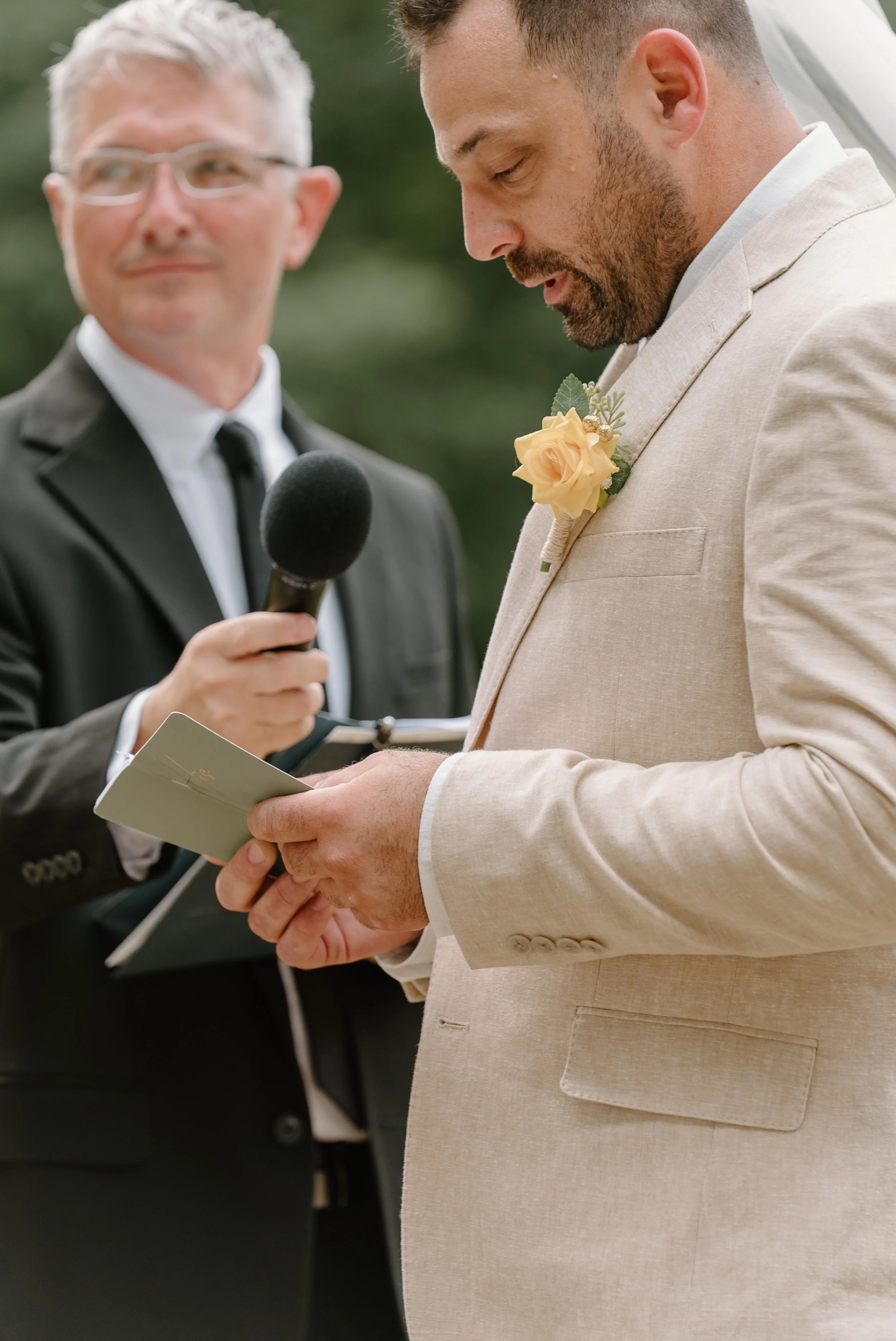 A man in a beige suit reads vows during a wedding ceremony, while an officiant in a black suit looks on for a wedding at Tunxis Country Club in Connecticut. 