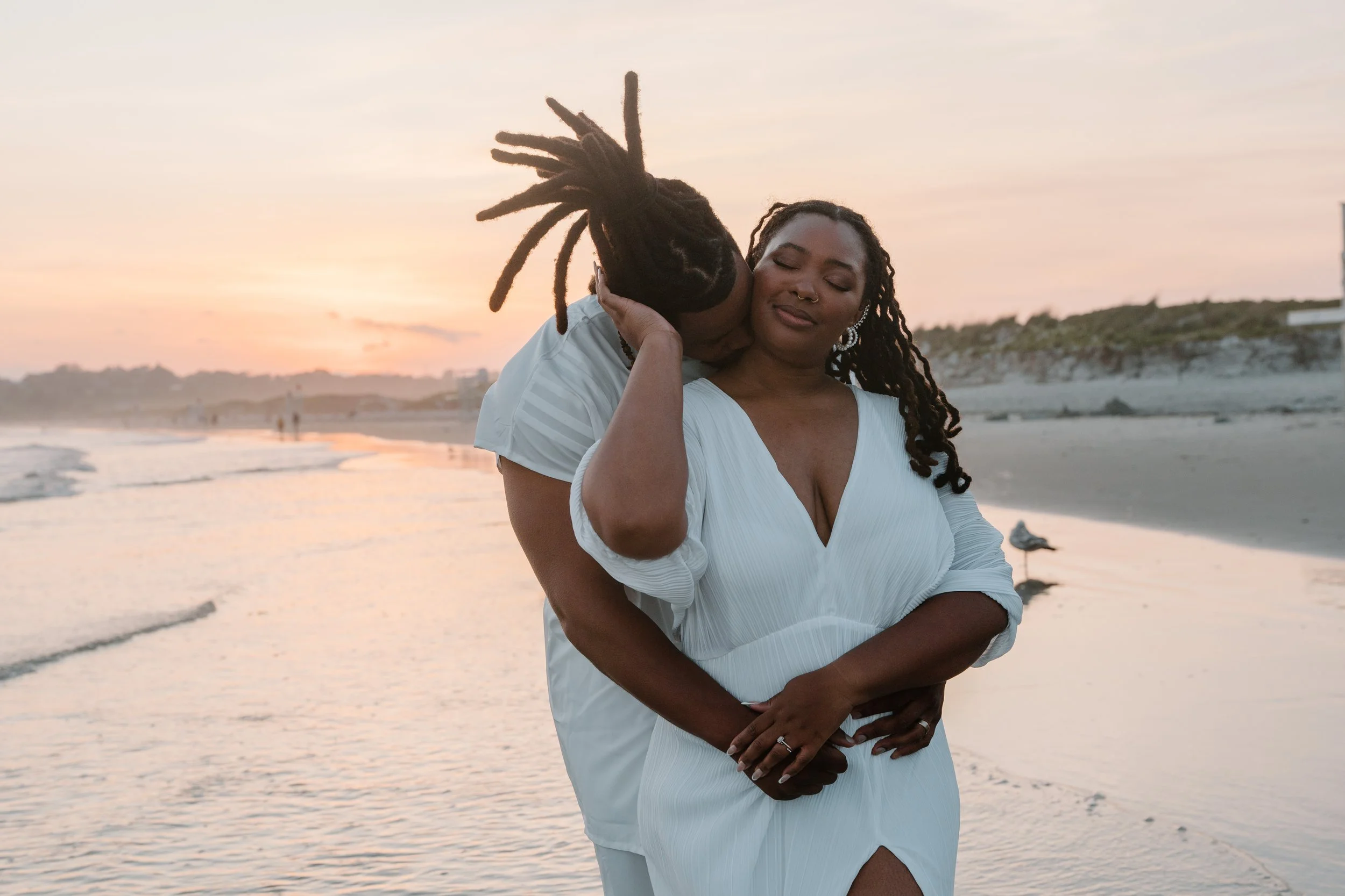 A couple embracing on the beach at sunset, with the man kissing the woman's cheek and the woman smiling with her eyes closed at an elopement at Second Beach in Rhode Island. 