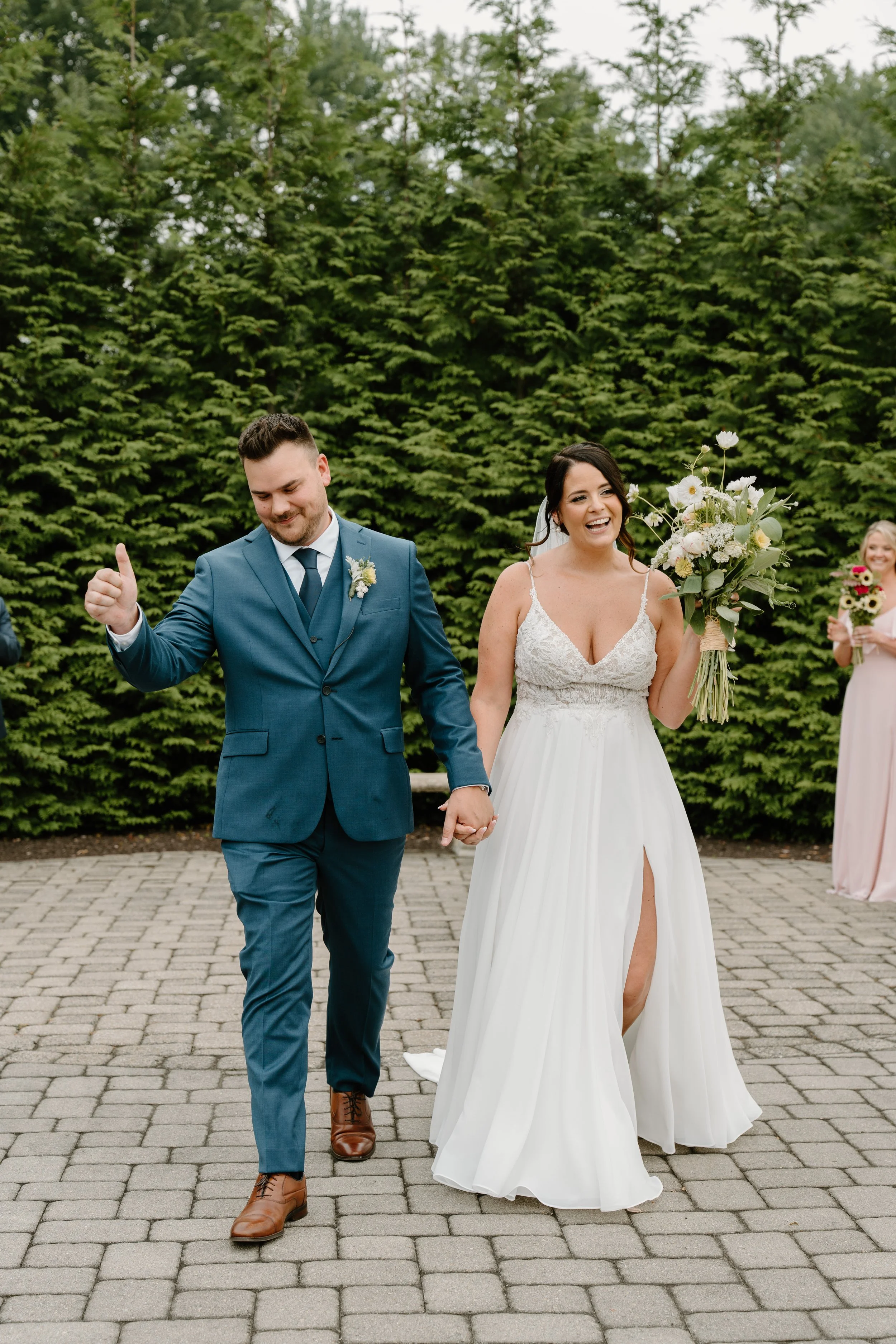 A newlywed couple holding hands and walking outdoors on a cobblestone path, with a lush green hedge in the background during a wedding at the Aqua Turf Club in Connecticut. 