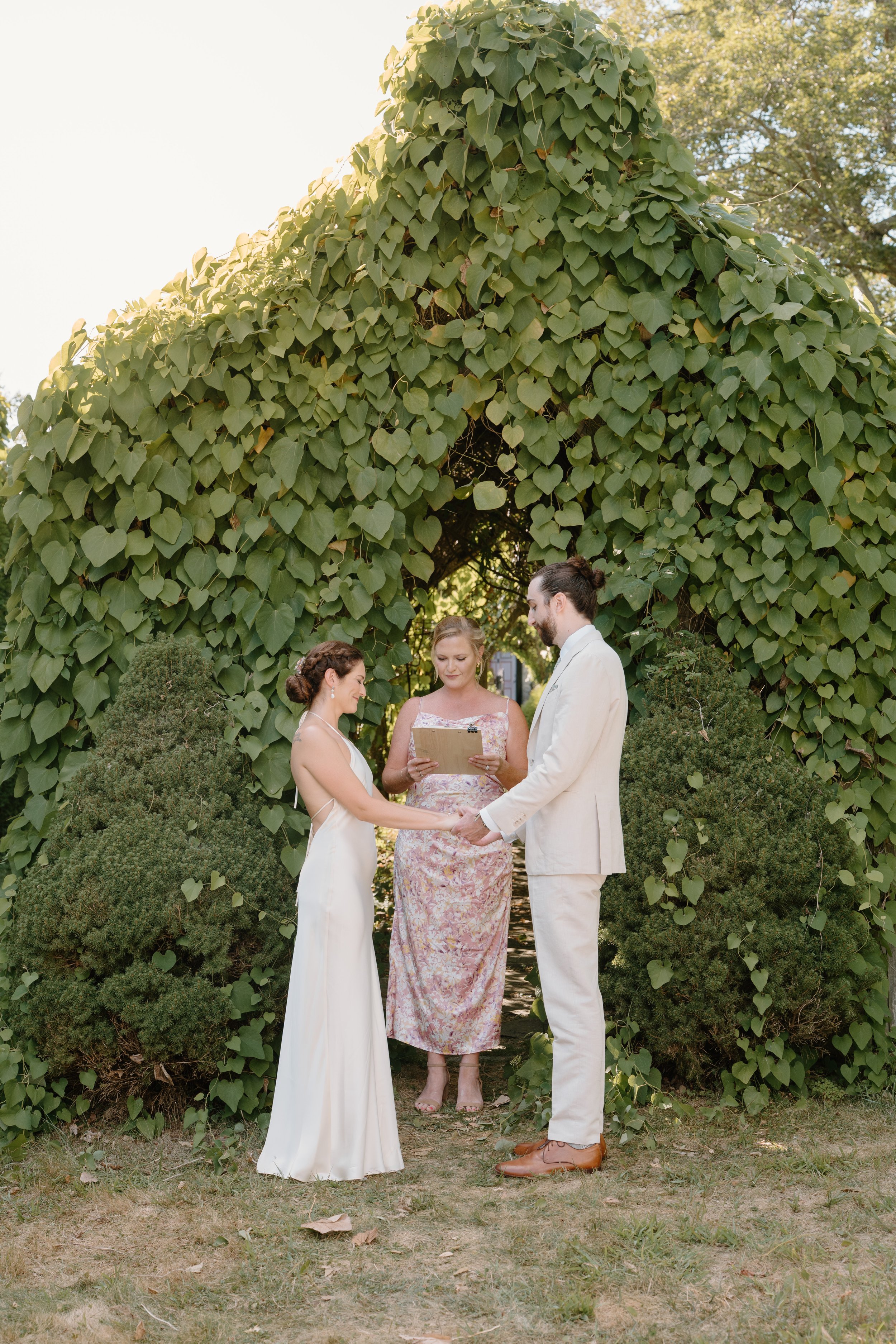 A couple getting married exchanging vows outdoors under a leafy arch, with an officiant standing between them, holding a tablet, in a garden setting at a wedding in Bath, Maine. 