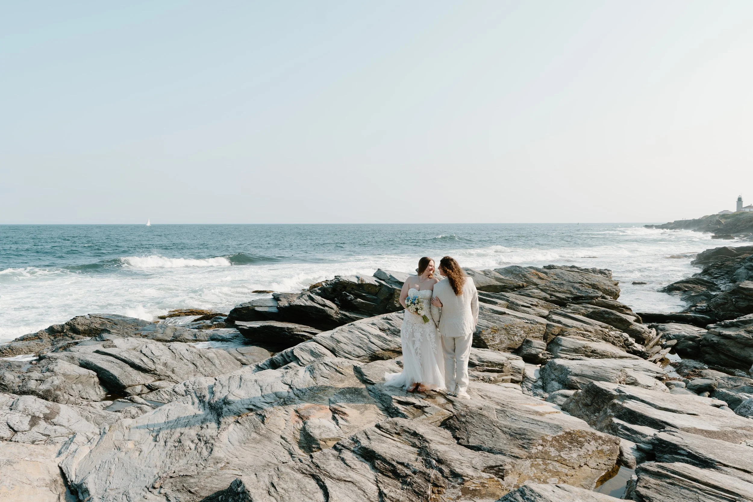 Two women in wedding attire standing on rocky shore near ocean, holding hands, with lighthouse in the background at an elopement at Beavertail State Park in Rhode Island. 