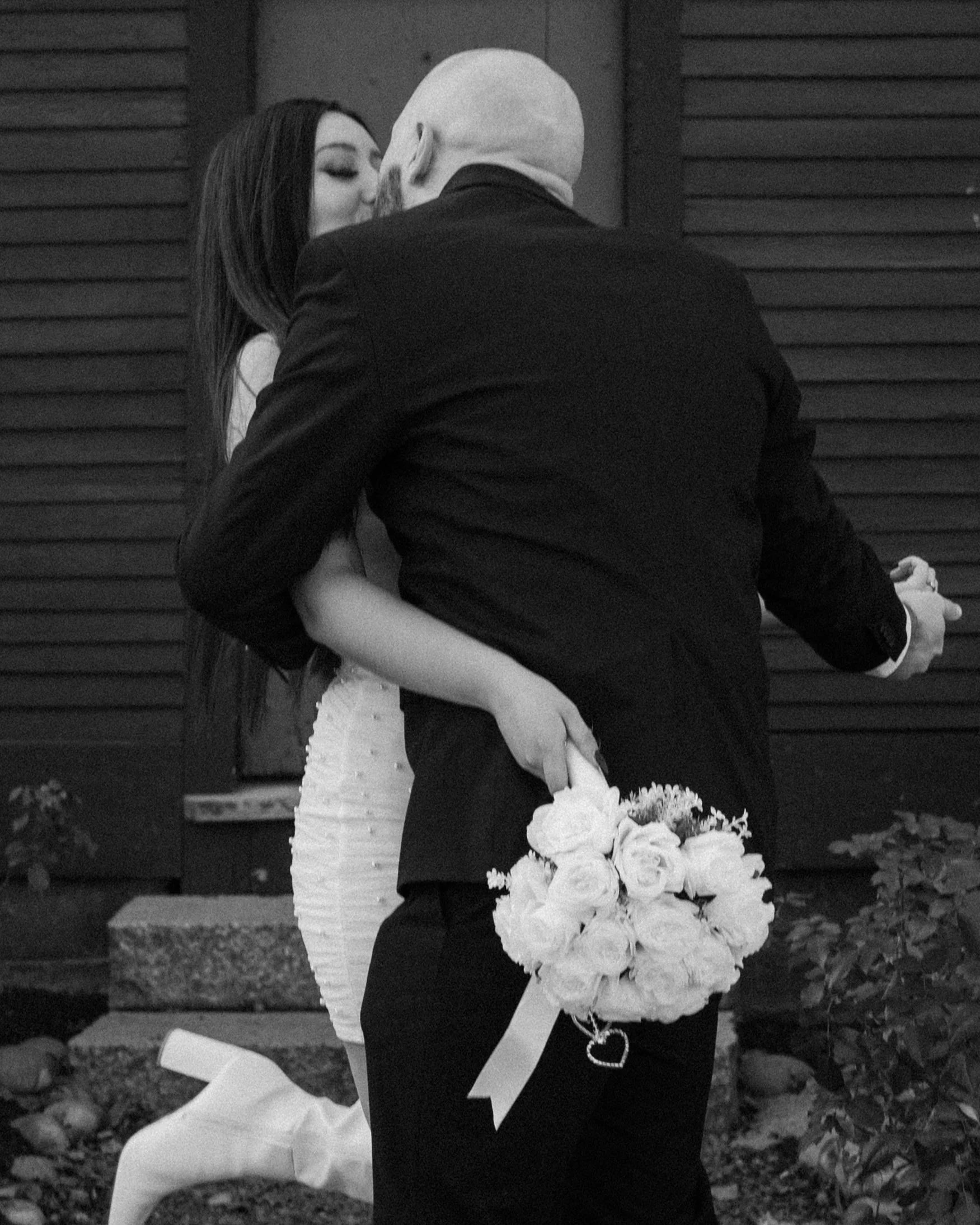 Black and white photo of a couple kissing, with the man holding a bouquet of flowers behind his back."