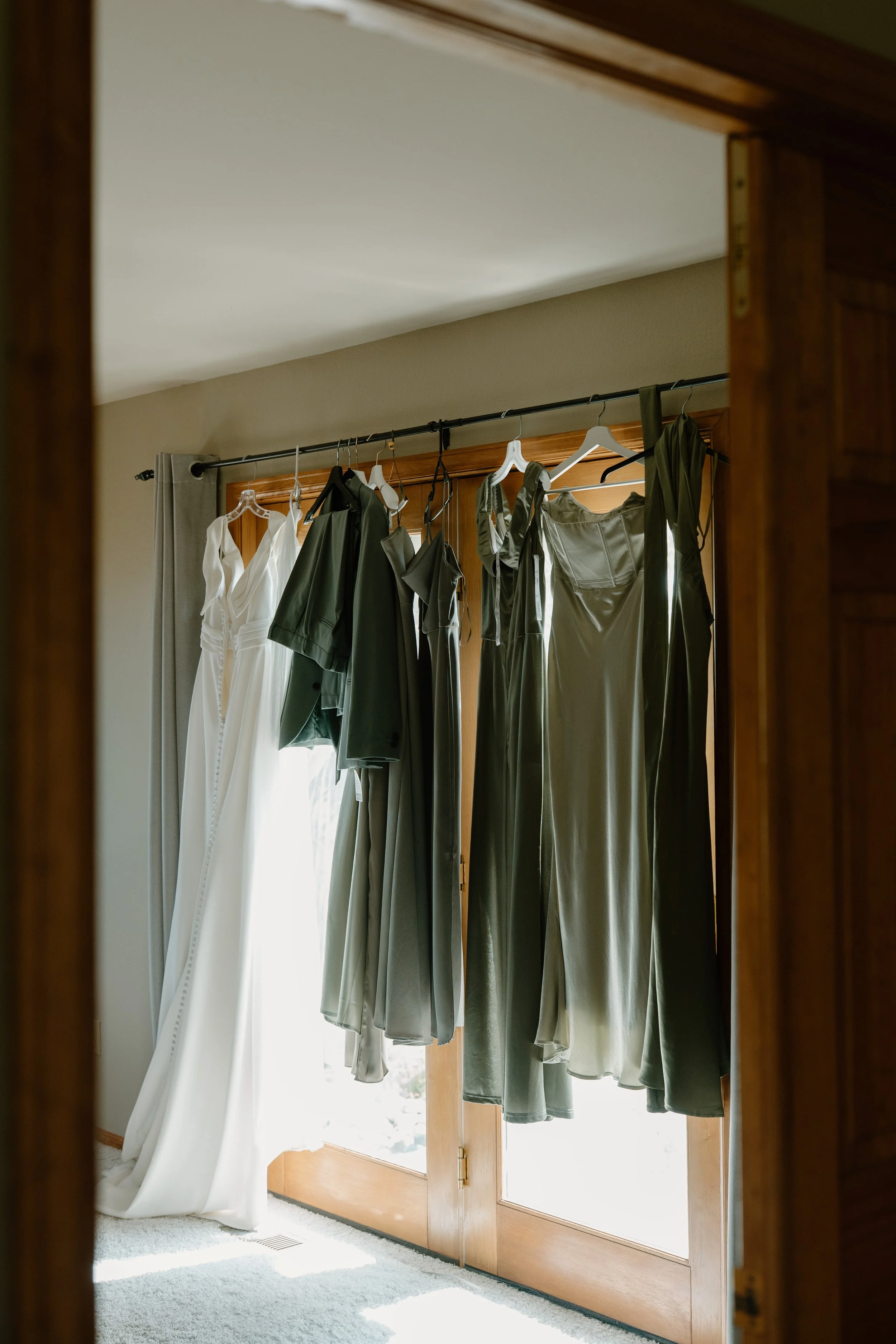 A collection of wedding dresses and bridesmaid dresses hanging on a rack in front of a wooden glass door at a wedding at Everett Ranch in Salida, Colorado.