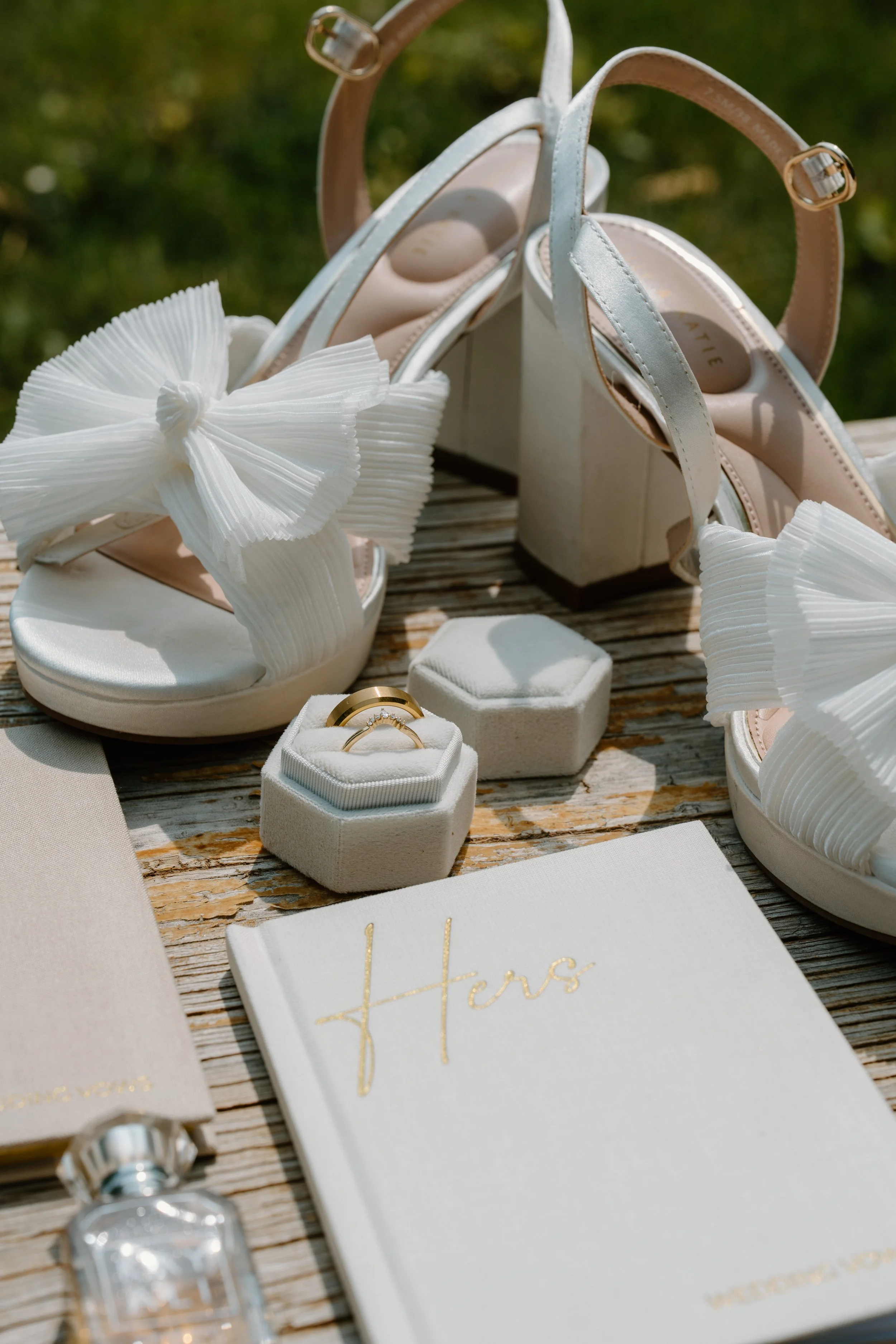 Wedding shoes, rings in a ring box, a guest book with 'Hers' written in gold, and other wedding accessories on a wooden surface at a wedding at Everett Ranch in Salida, Colorado.