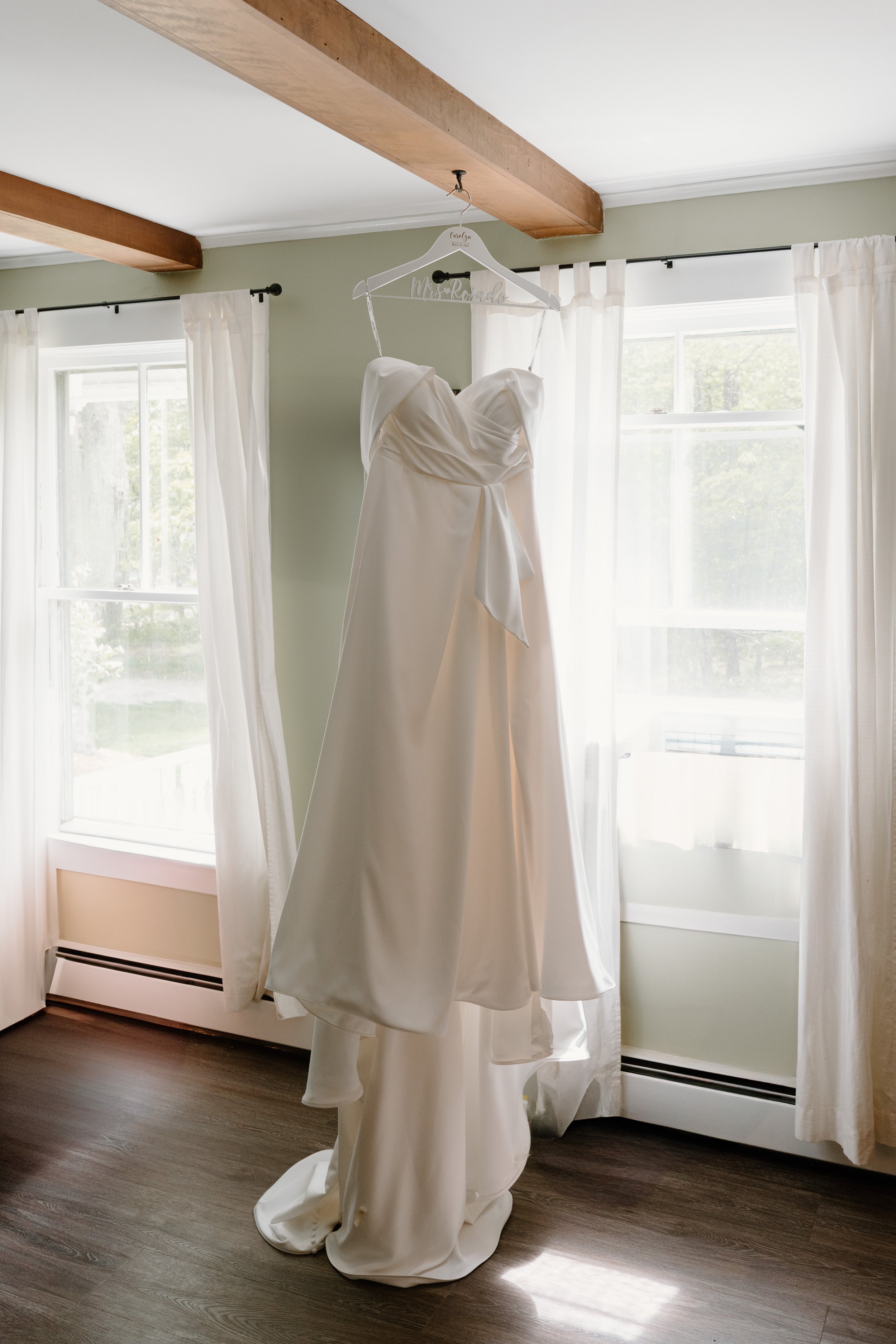 White wedding dress hanging on a hanger in a room with two windows and white curtains for a wedding at Holiday Hill Day Camp in Connecticut.