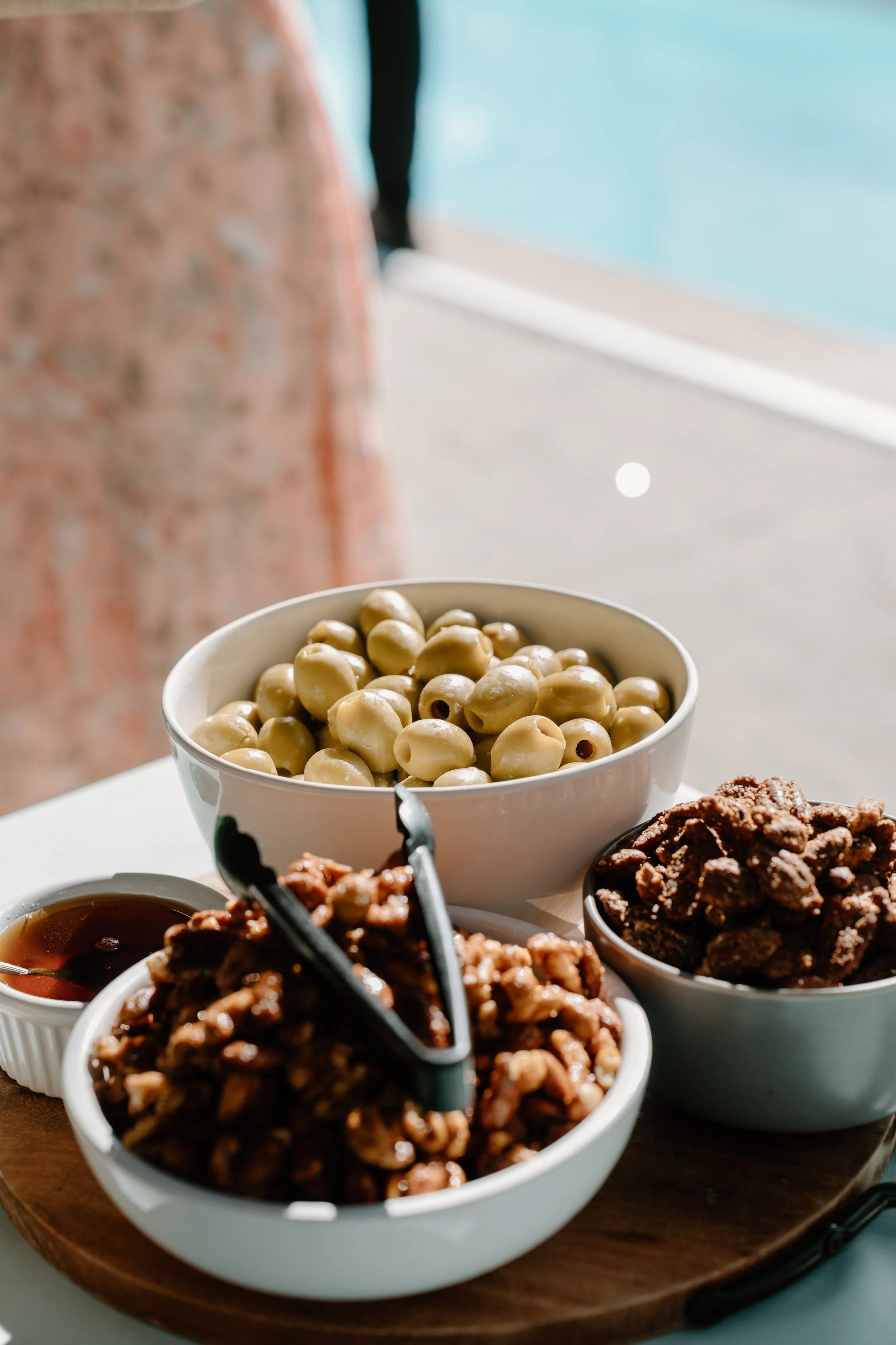 Bowls of white and dark chocolate-covered nuts with a small bowl of syrup, placed on a wooden tray near a swimming pool at a wedding in Bath, Maine. 
