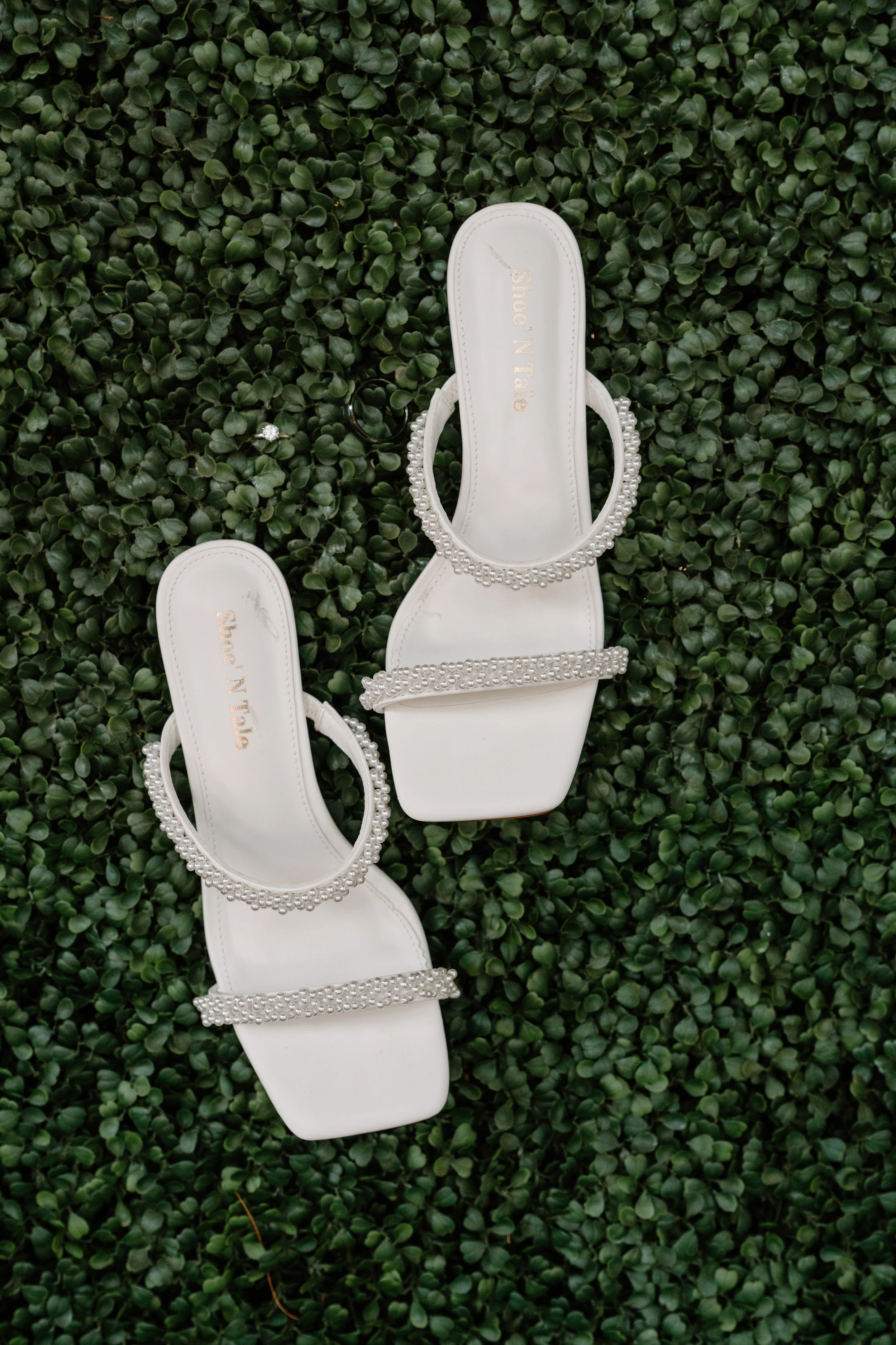 Pair of white sandals with jeweled straps on a green leafy surface for a wedding at Tunxis Country Club in Connecticut. 