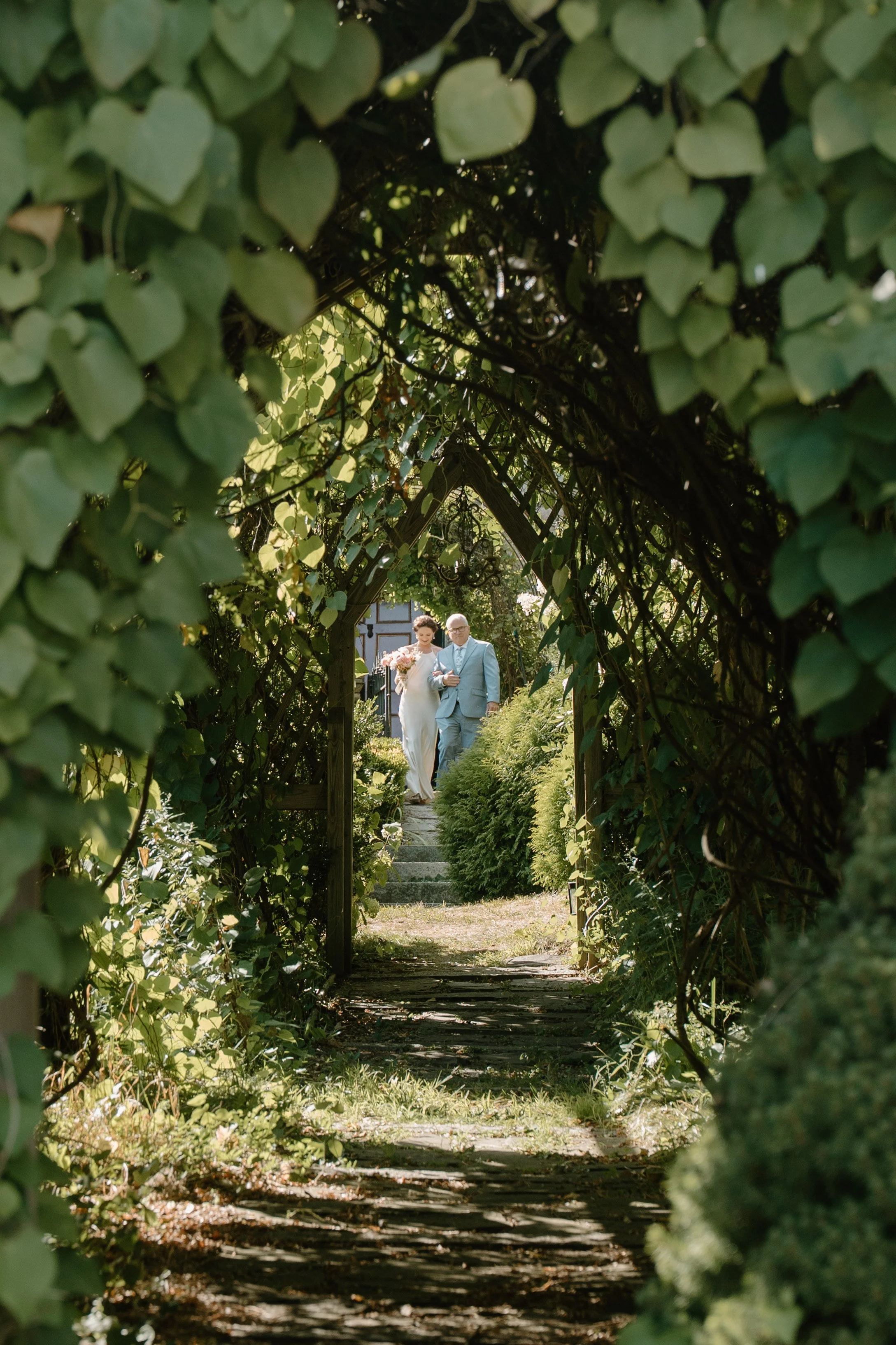A bride and dad walking through a garden archway, viewed through greenery and leaves at a wedding in Bath, Maine. 