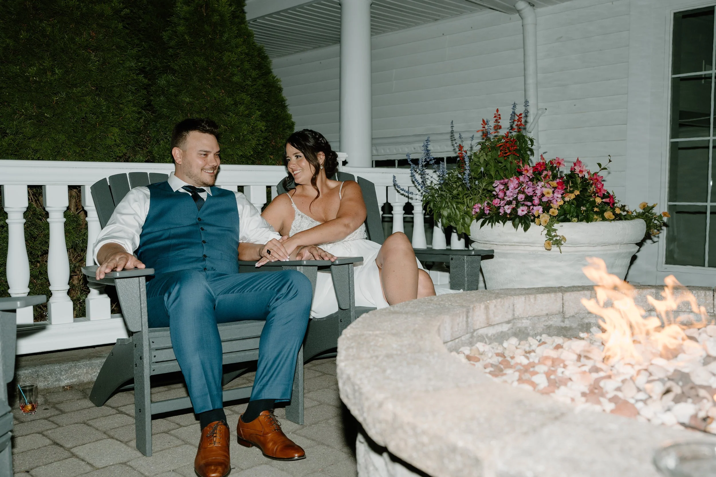 A couple dressed in wedding attire sitting on outdoor chairs near a fire pit, smiling at each other. There are colorful flowers in large pots behind them and green bushes in the background during a wedding at the Aqua Turf Club in Connecticut. 