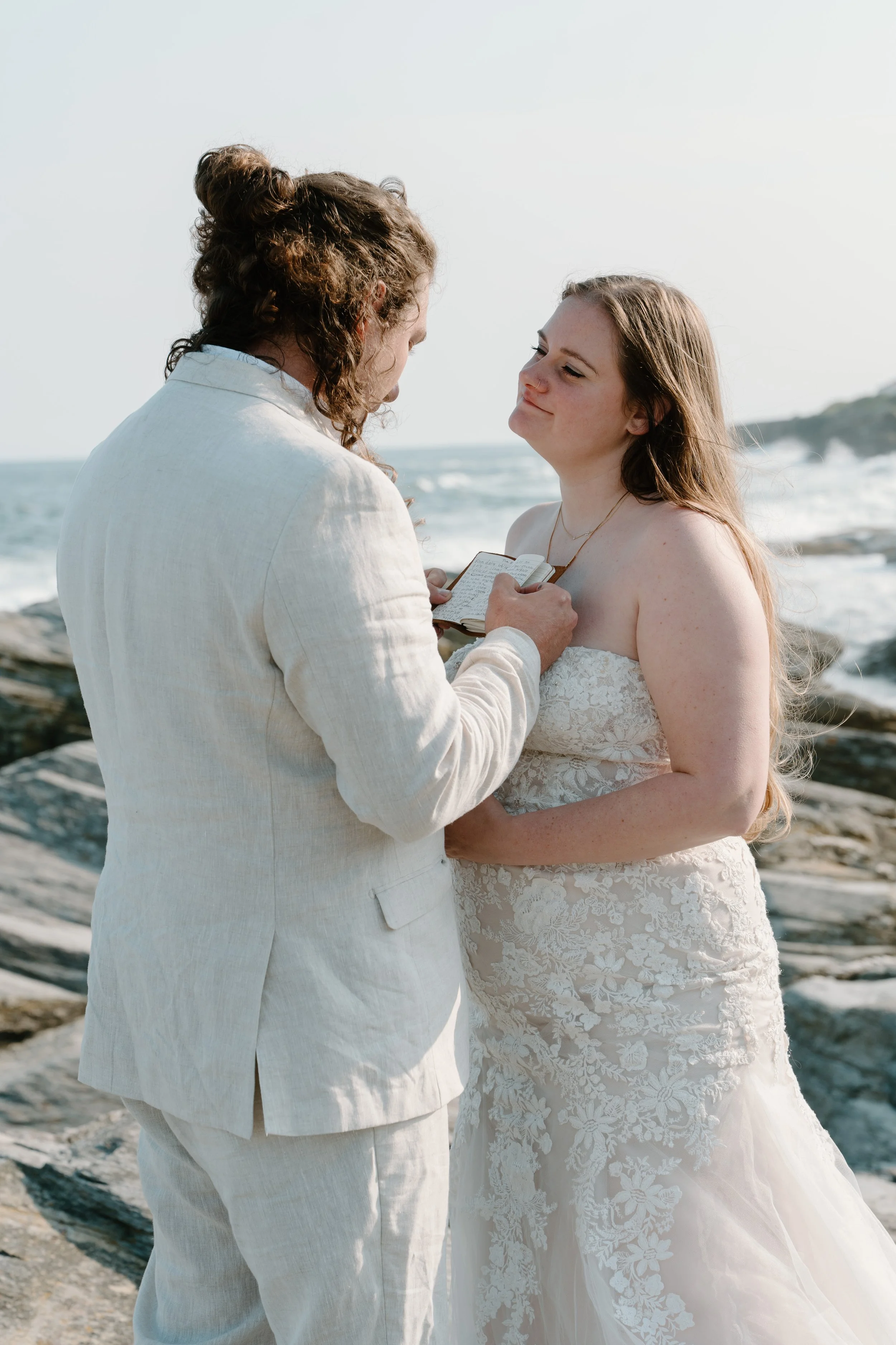 A couple getting married outdoors near the ocean, with one person reading vows from a small book at an elopement at Beavertail State Park in Rhode Island. 
