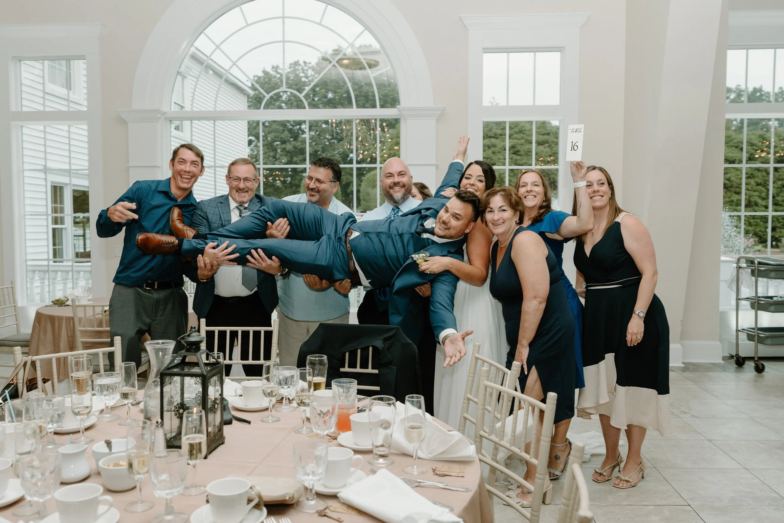 Group of men and women at a wedding reception lifting a groom in a blue suit in a banquet hall with large windows during a wedding at the Aqua Turf Club in Connecticut. 