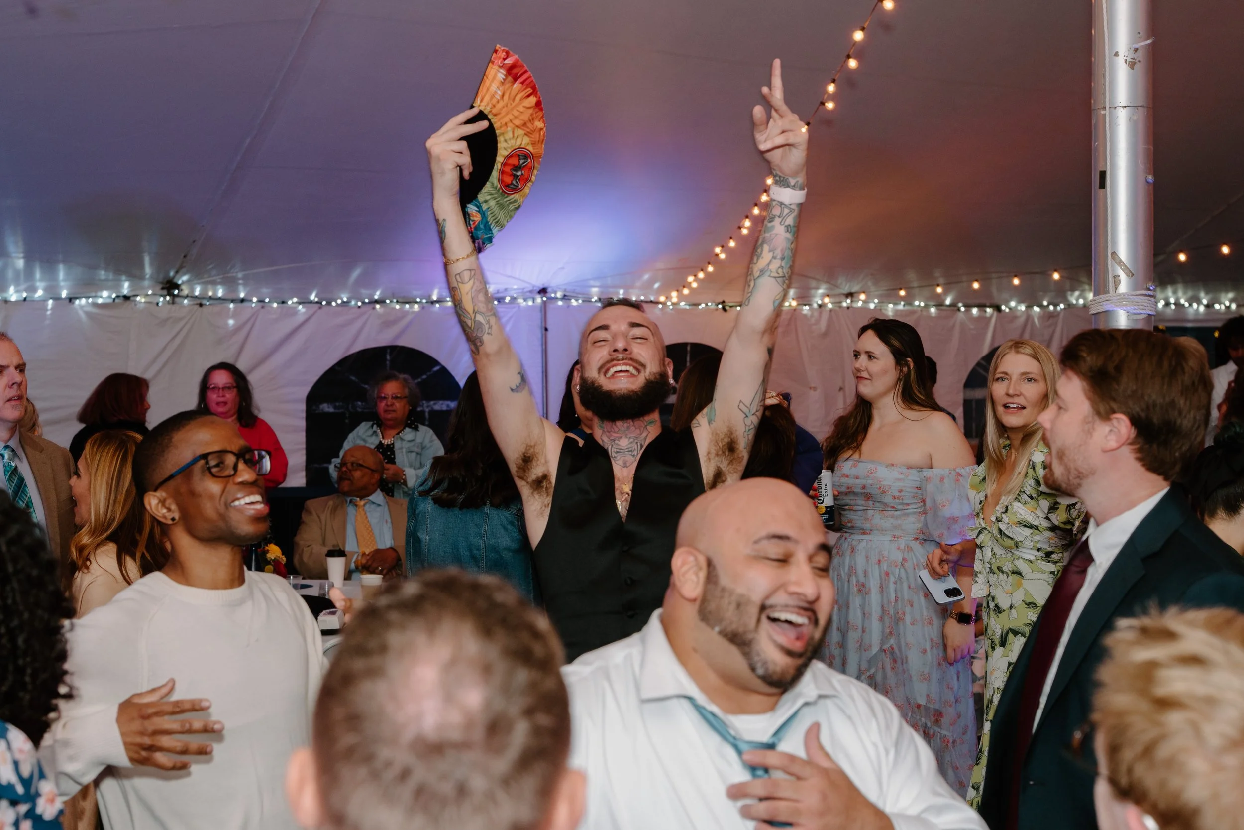 A group of people at a celebration under a tent with string lights. A man in the center with tattoos, a beard, and a sleeveless shirt is smiling, raising his arms, and holding a colorful fan for a wedding at Holiday Hill Day Camp in Connecticut.