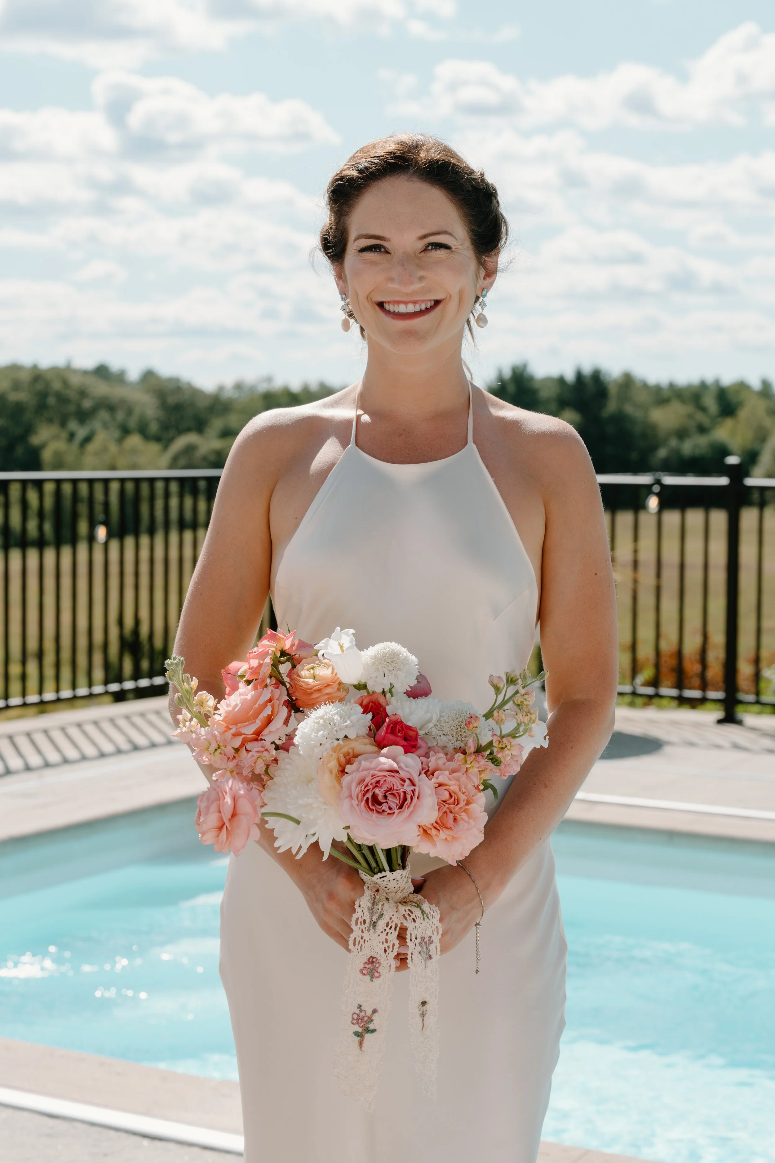 A woman in a white dress holding a bouquet of pink, white, and red flowers outdoors near a pool, smiling at the camera on a sunny day with a partly cloudy sky at a wedding in Bath, Maine. 