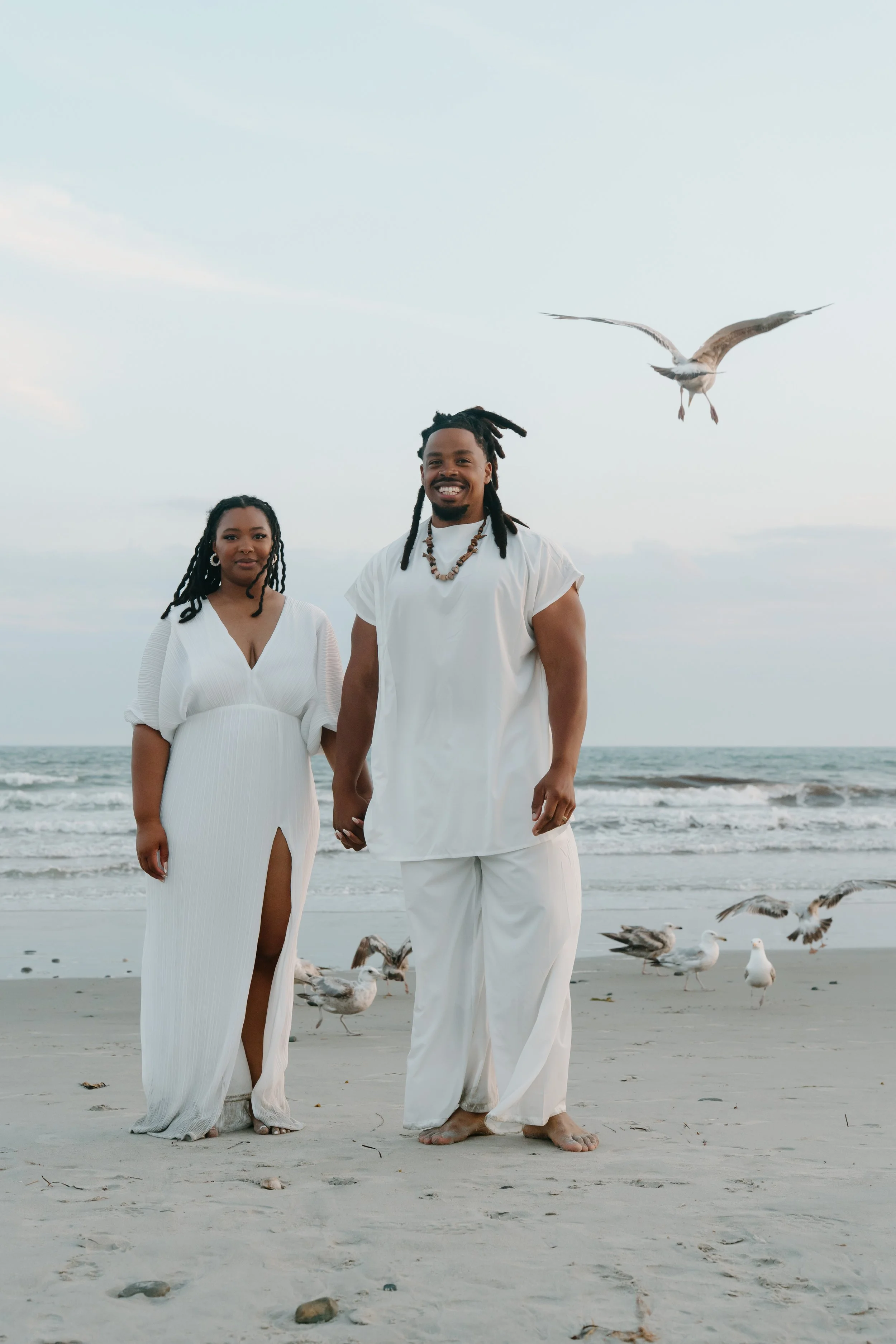 A couple standing on the beach holding hands, dressed in white, with seagulls flying around them at an elopement at Second Beach in Rhode Island. 