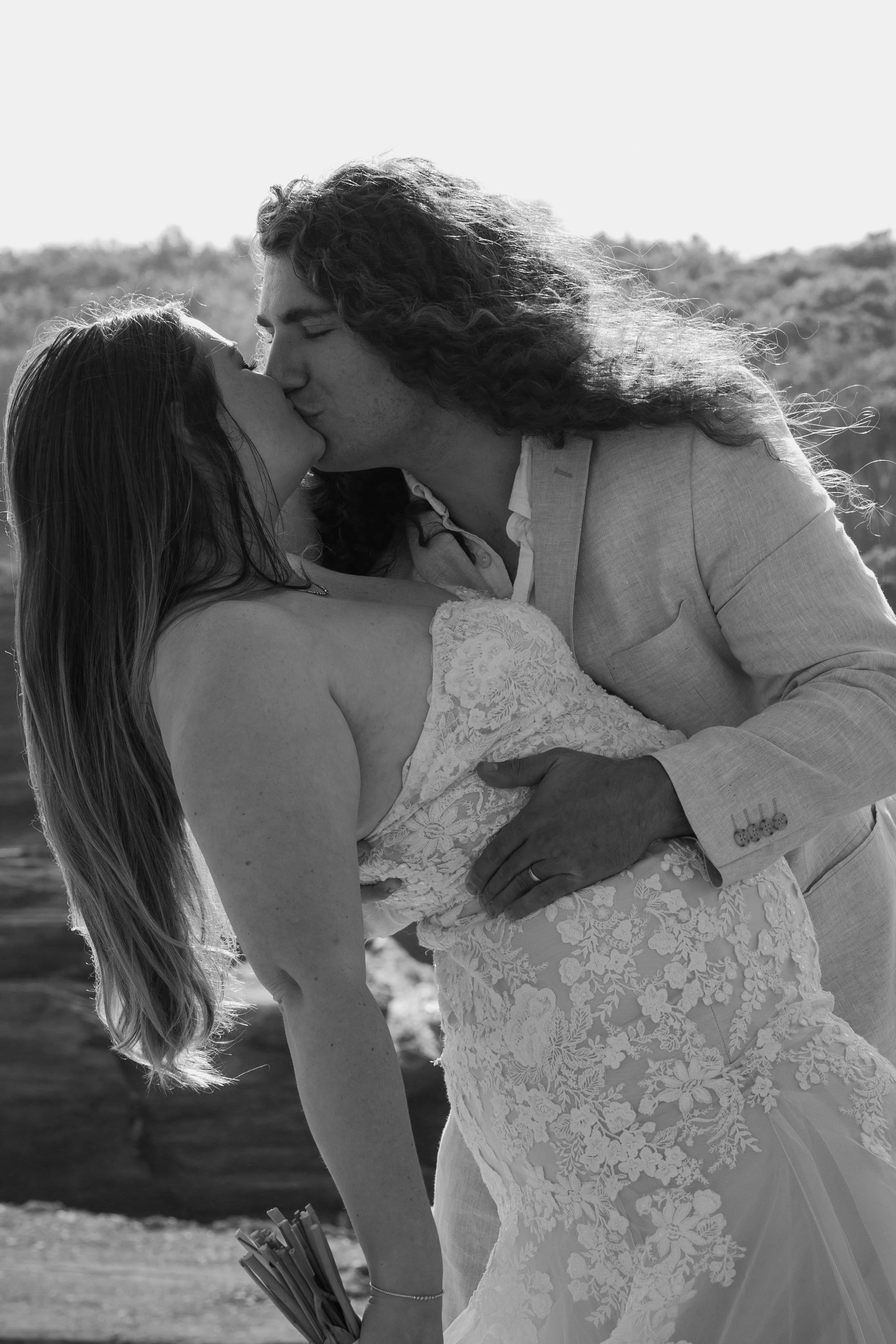 Black and white photo of a couple kissing outdoors, with the woman in a lace dress and holding a bouquet, and the man in a light-colored suit at an elopement at Beavertail State Park in Rhode Island. 