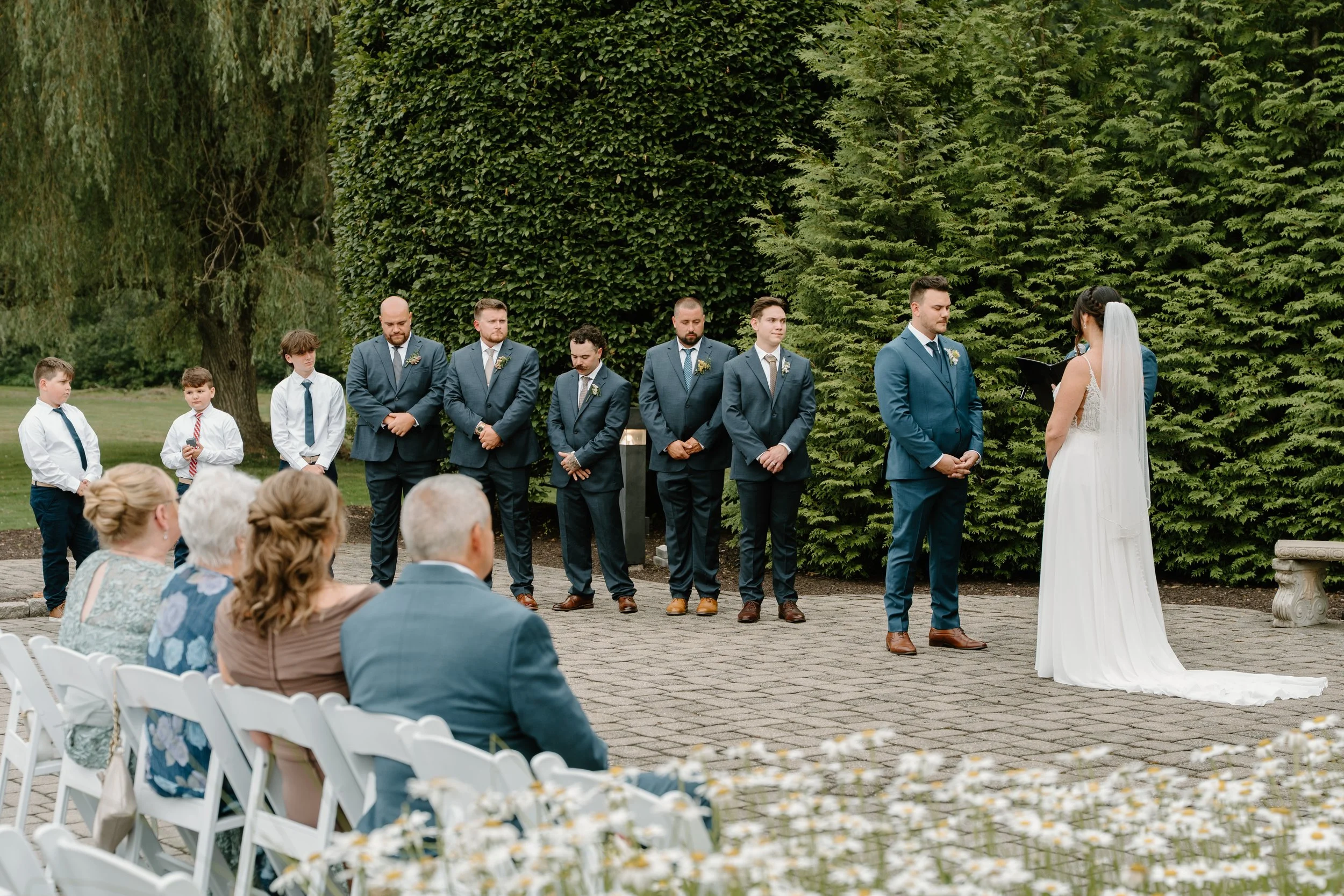 A wedding ceremony taking place outdoors with a bride, groom, and groomsmen standing in line, and guests seated in white chairs, surrounded by green trees and bushes during a wedding at the Aqua Turf Club in Connecticut. 