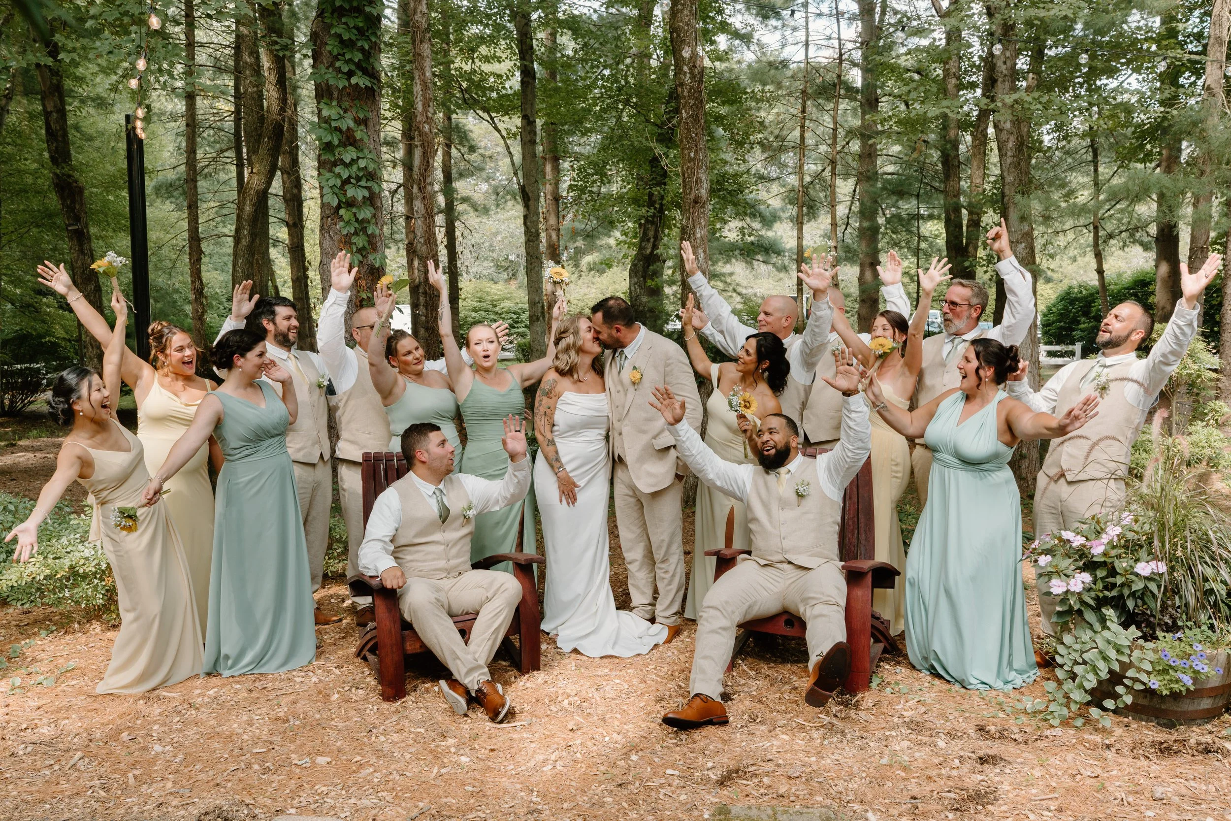 A wedding party celebrating outdoors in a wooded area, with the bride and groom kissing in the center, surrounded by bridesmaids and groomsmen with arms raised and smiling for a wedding at Tunxis Country Club in Connecticut. 