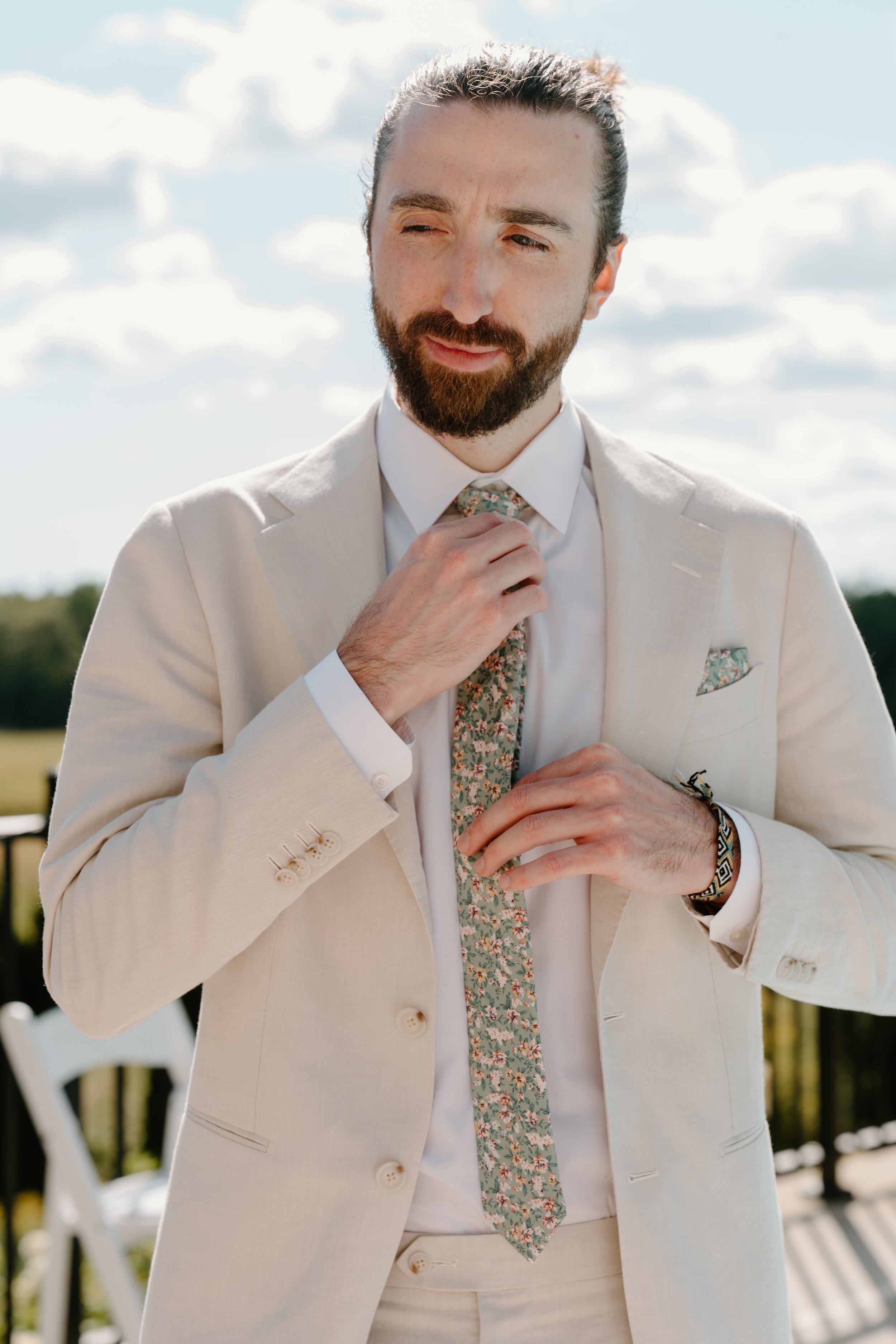 Man adjusting floral patterned tie while wearing a cream suit outdoors on a sunny day at a wedding in Bath, Maine. 