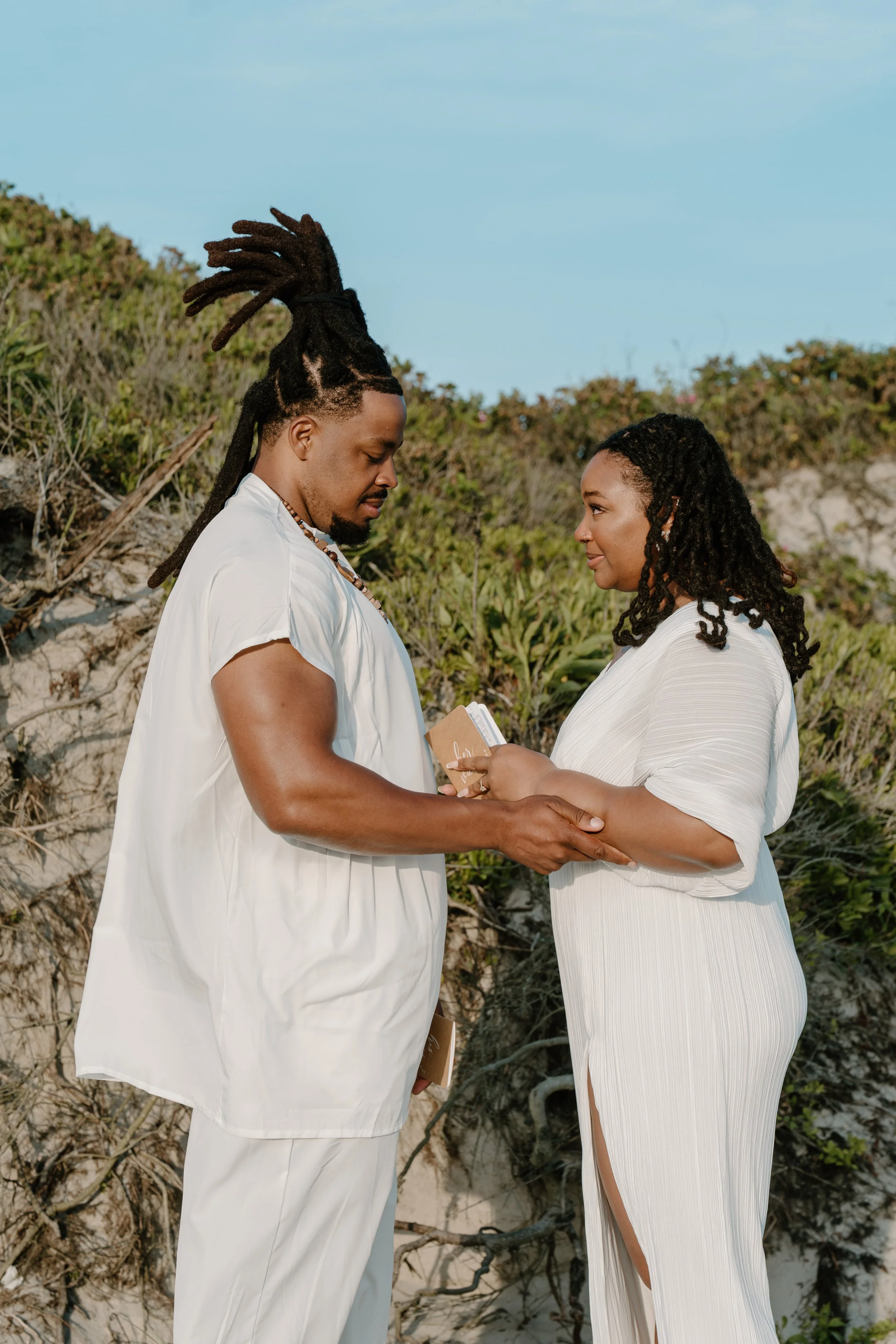 A couple dressed in white holding hands and exchanging vows outdoors on a beach with sand dunes and green bushes in the background at an elopement at Second Beach in Rhode Island. 