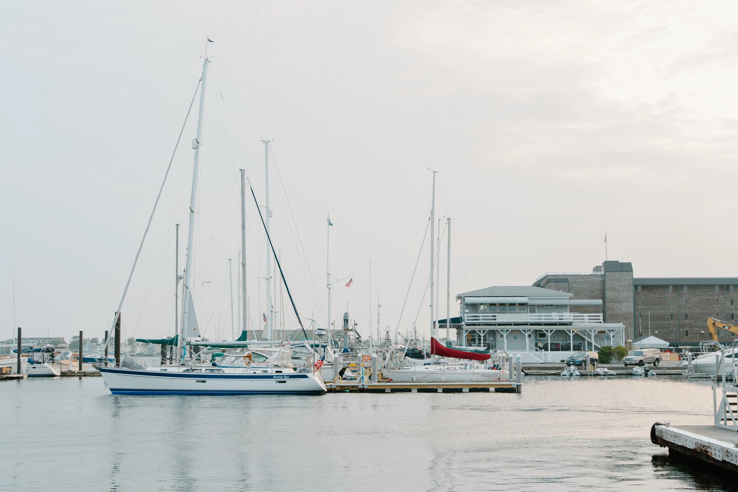 Marina with sailboats docked, a waterfront building, and parked cars in Newport, Rhode Island. 