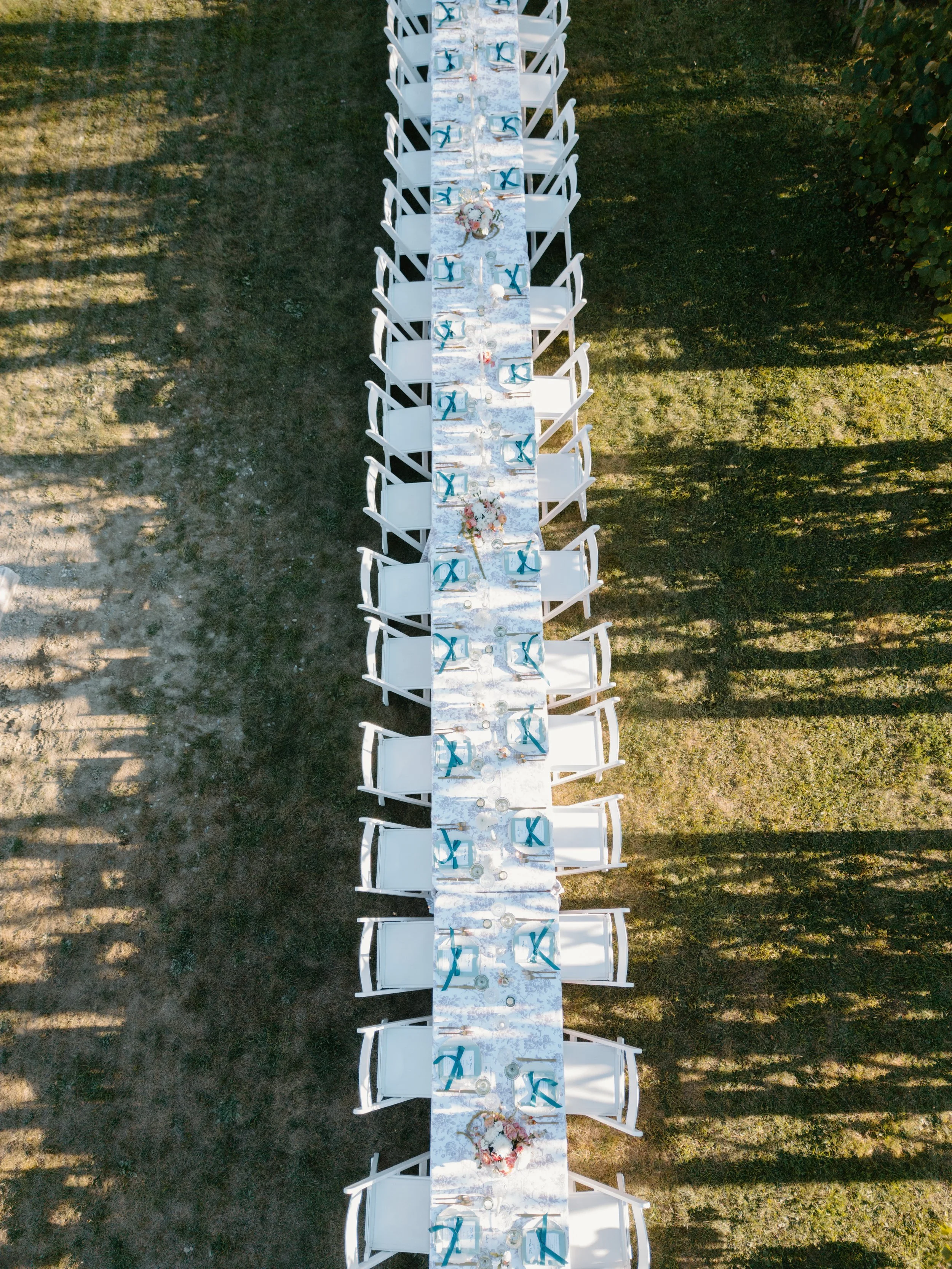 Overhead view of a long outdoor dinner table set with white chairs, decorated with floral centerpieces, blue napkins, and glassware, on a grassy lawn at a wedding in Bath, Maine. 